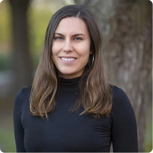 A woman with long brown hair, wearing a black turtleneck, smiling outdoors in front of a tree and greenery.