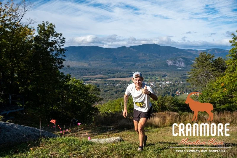 A smiling man in running gear with a white shirt and shorts, giving a thumbs-up while running outdoors on a trail in a mountainous area of North Conway, New Hampshire, during daytime with a scenic view of mountains and blue sky.