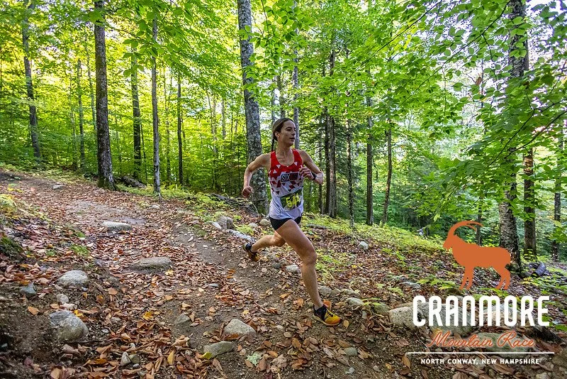A female trail runner in athletic gear running on a rocky dirt trail through a forest of green trees. The image features a logo of a goat and the text ‘Crankmore Mountain Race’ with location details ‘North Conway, New Hampshire’.