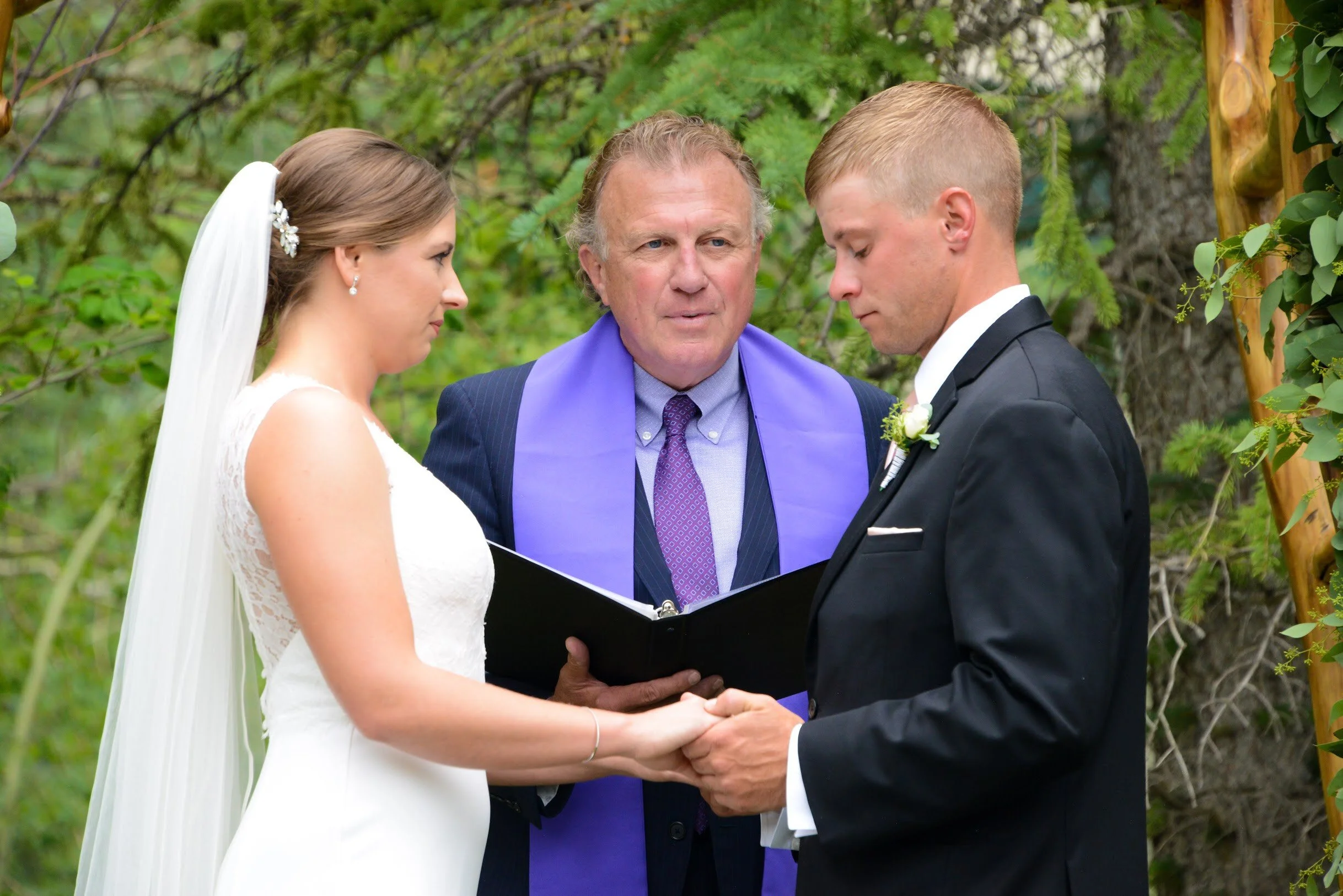 A bride and groom hold hands during their wedding ceremony outdoors, with a officiant standing behind them, surrounded by greenery.