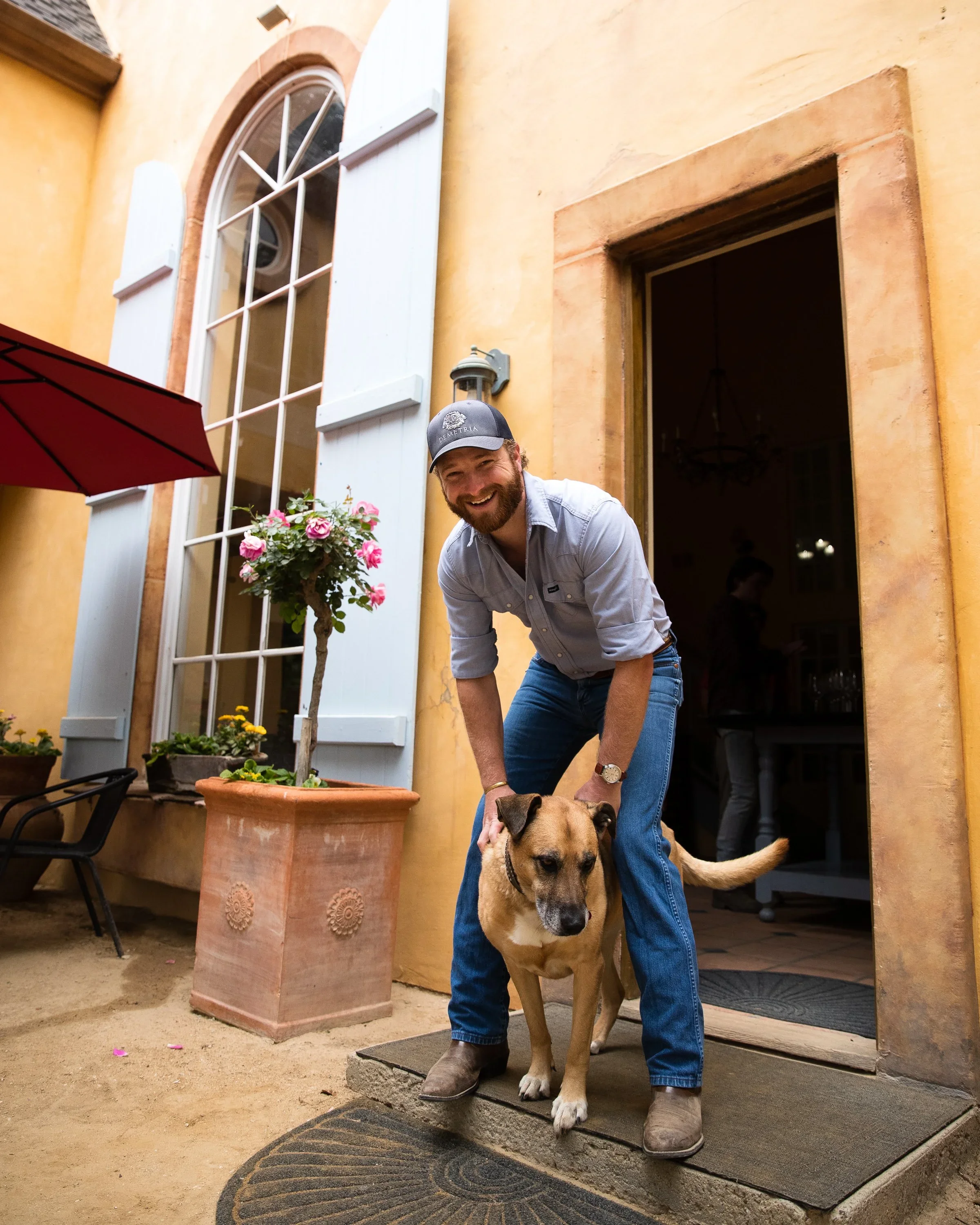 A man with a beard, wearing a cap and casual clothes, is smiling and bending down to pet a large brown dog just outside the doorway to a yellow stucco house with light blue shutters and a large arched window.