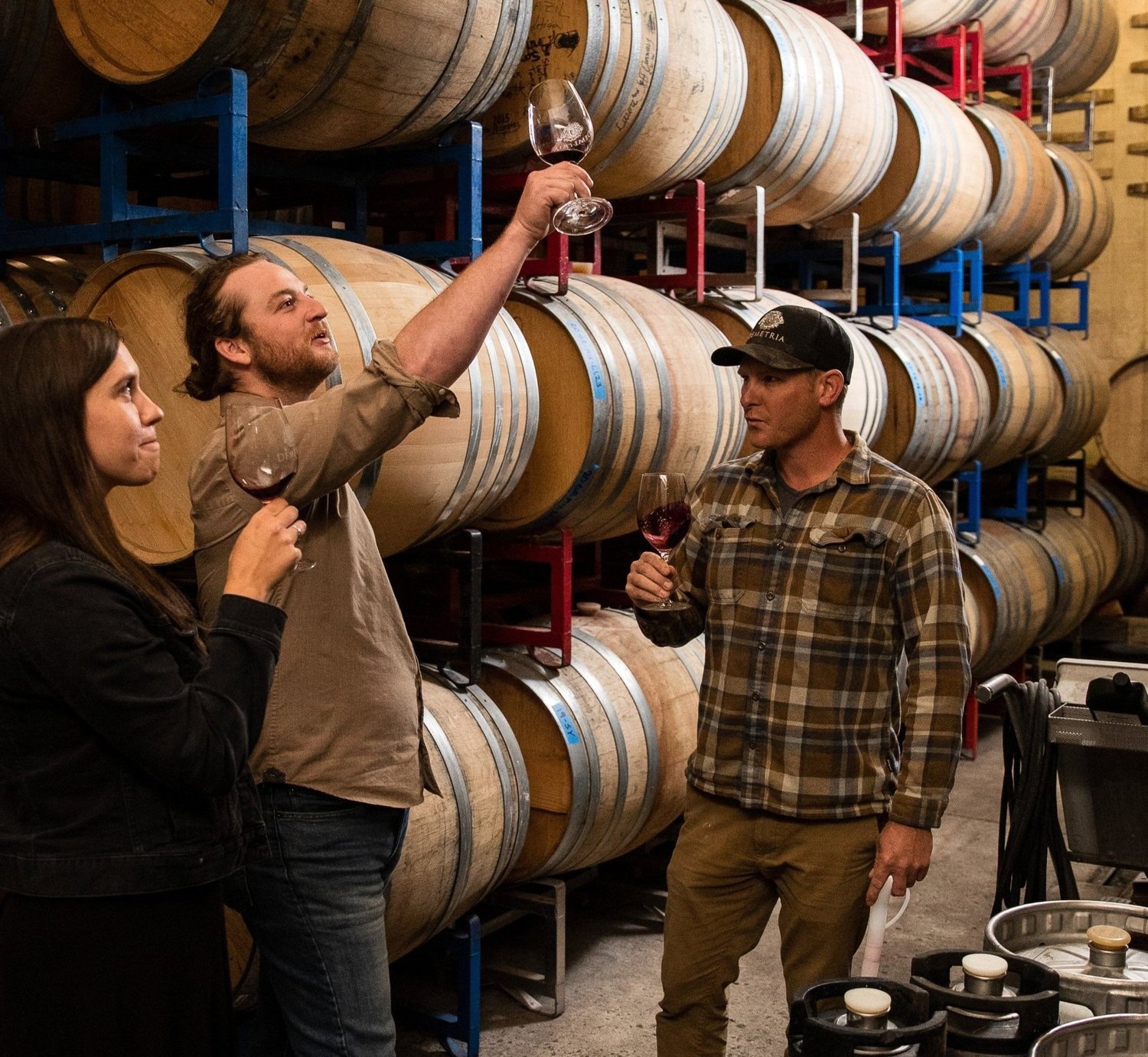 Three people tasting wine in a winery, with large oak barrels stacked in the background. Two men and one woman, with the man in the middle holding up a glass of red wine.