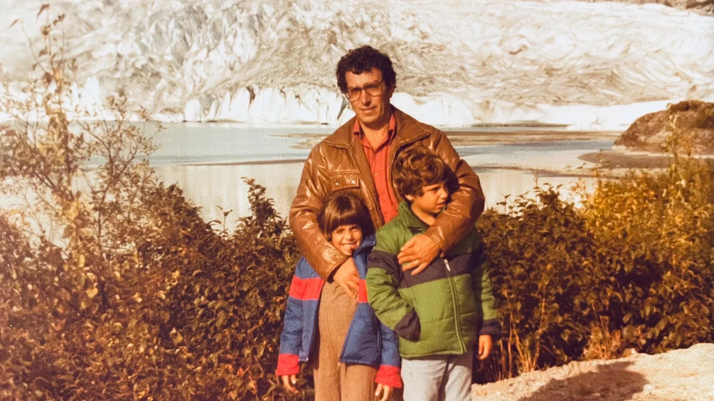 A man with two children standing outdoors near a body of water and snowy cliffs, with bushes in the foreground. The man is wearing a brown leather jacket and glasses, the girl has a colorful jacket, and the boy is in a green and black jacket.
