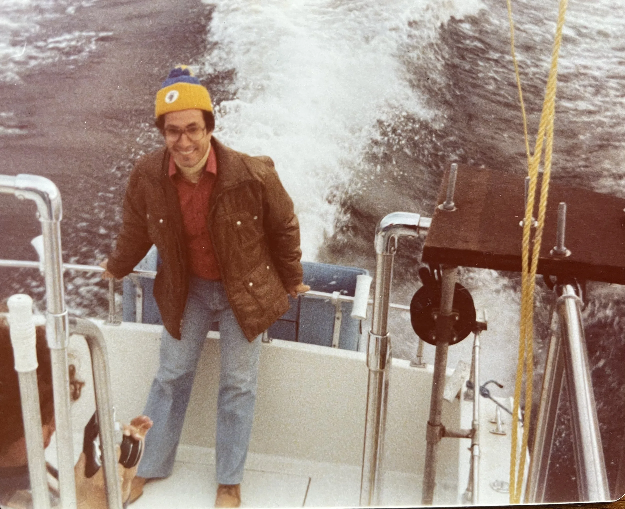A man with glasses, a yellow hat, brown jacket, and red shirt is standing on a boat, smiling at the camera. The boat is moving through water, creating waves behind him.