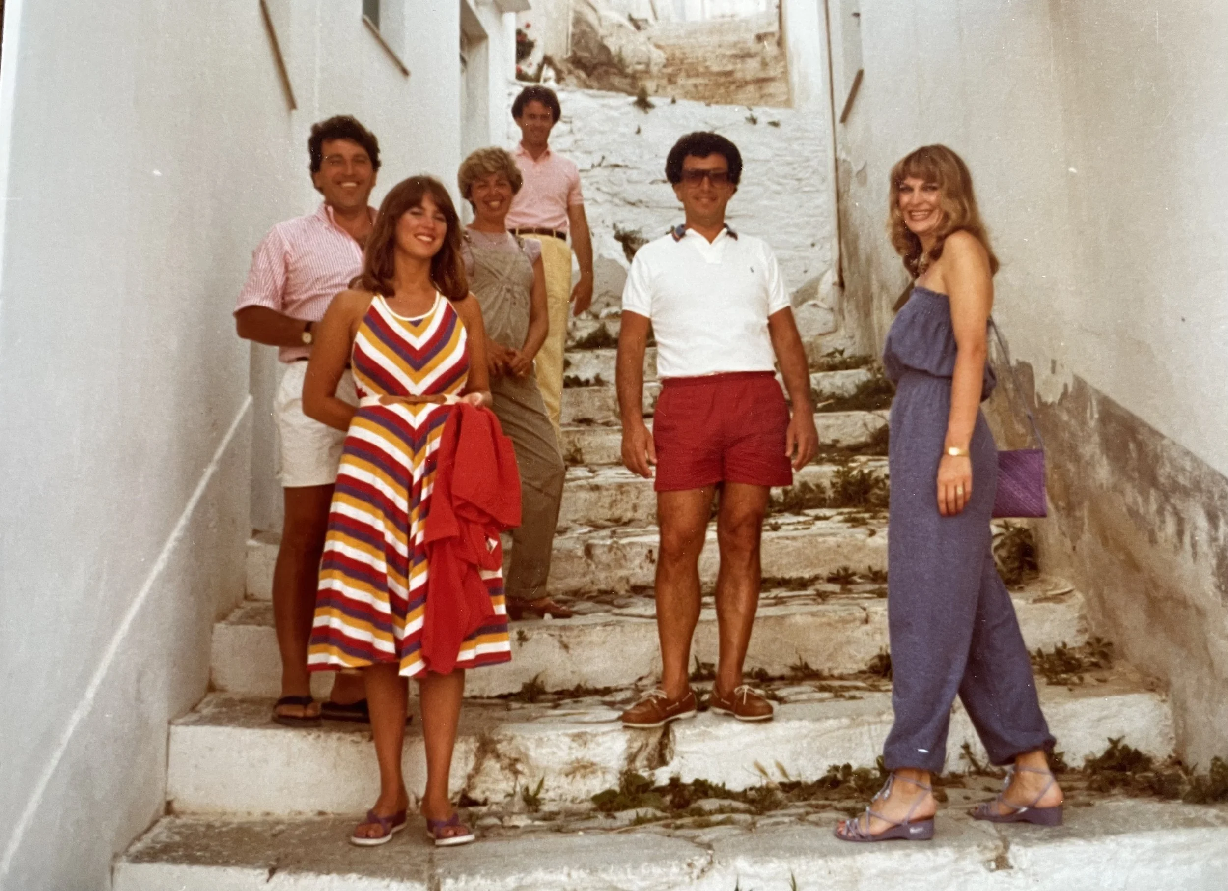 Group of seven people posing on stone stairs in an outdoor setting, dressed in casual summer clothing, with a rocky hillside in the background.