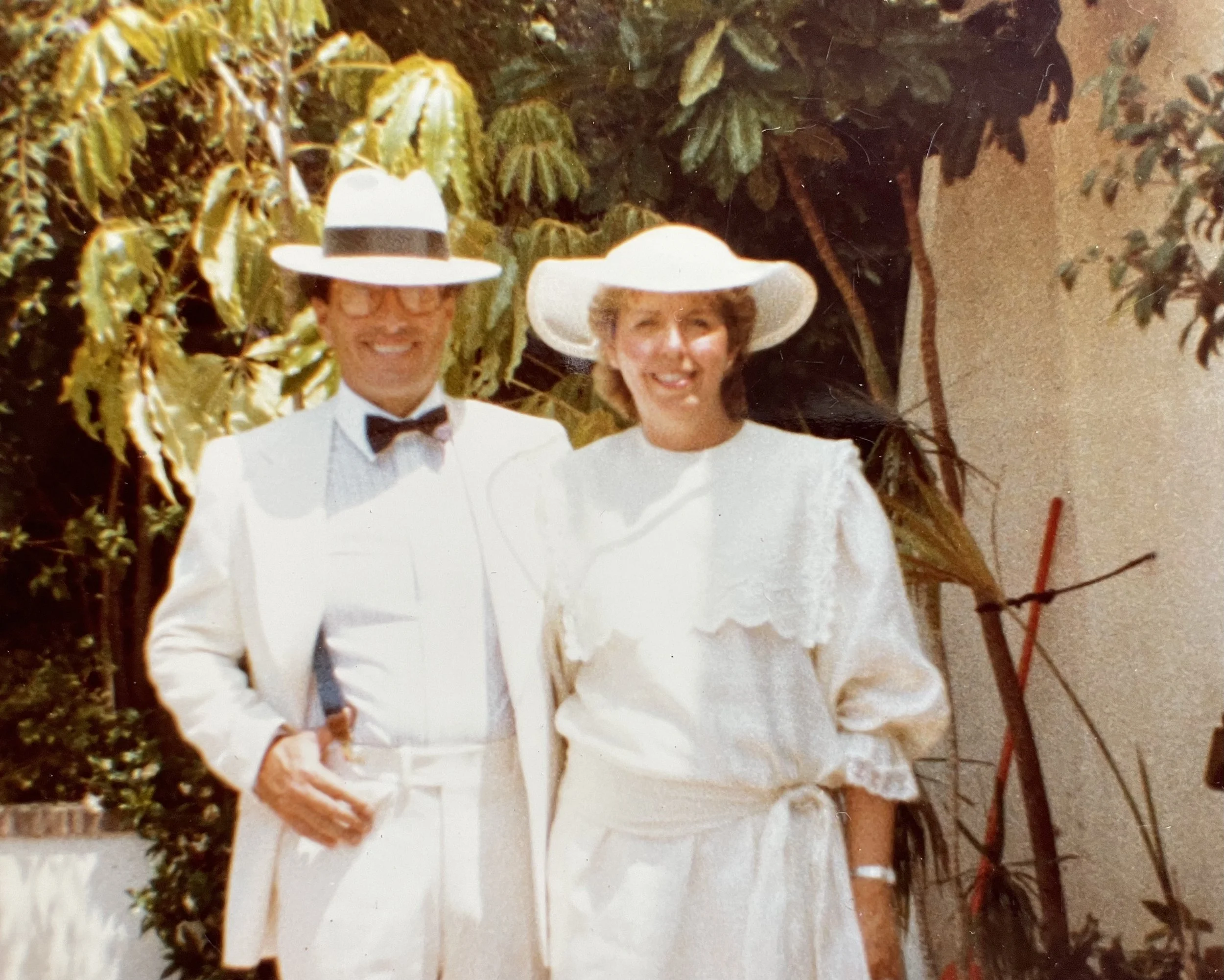 A man and woman dressed in white at a sunny outdoor event, with tropical plants in the background, both wearing white hats and smiling.