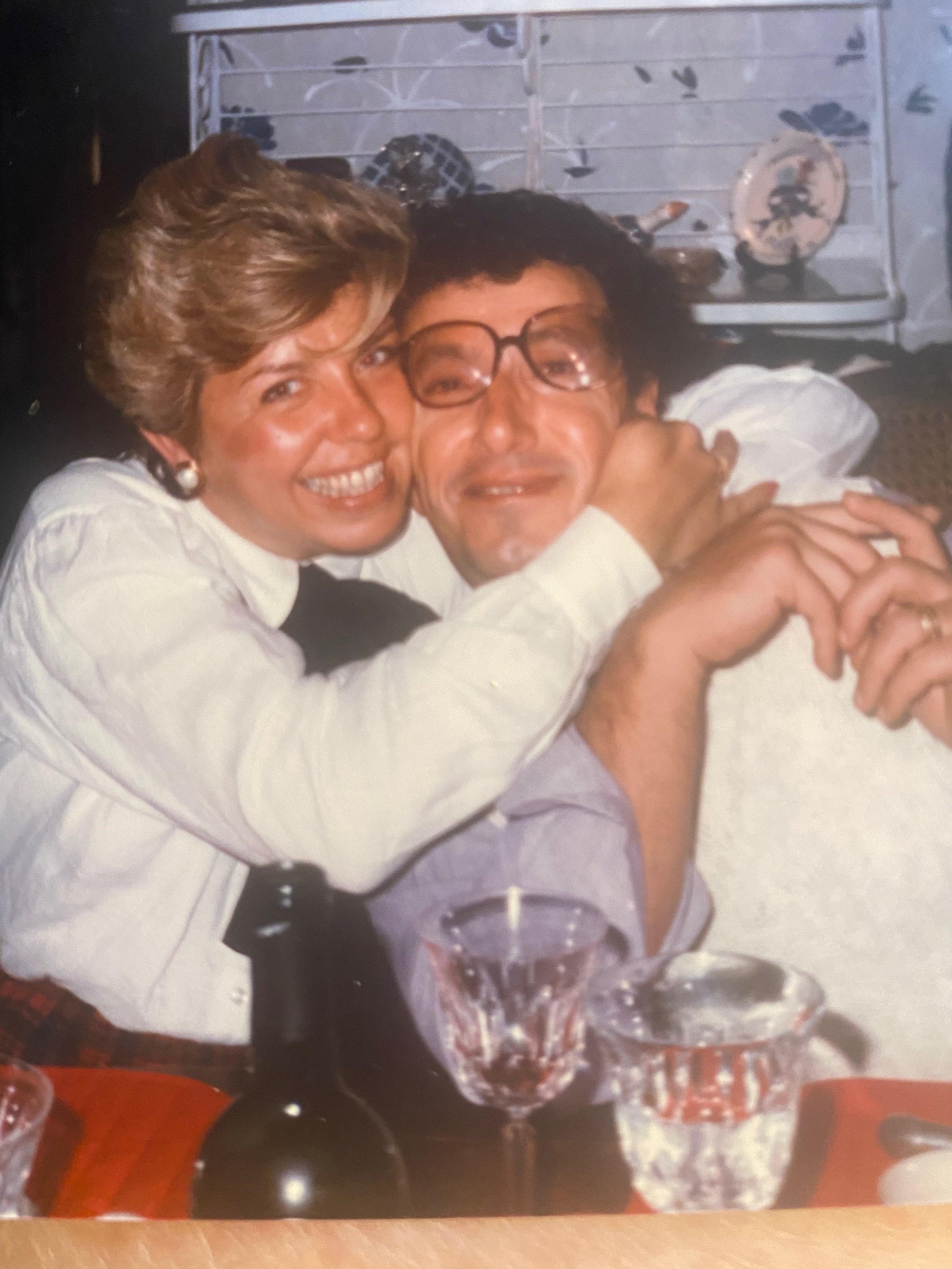 A woman with short hair and pearl earrings hugging a man with glasses and curly hair, both smiling at the camera, at a table with glasses and a bottle.