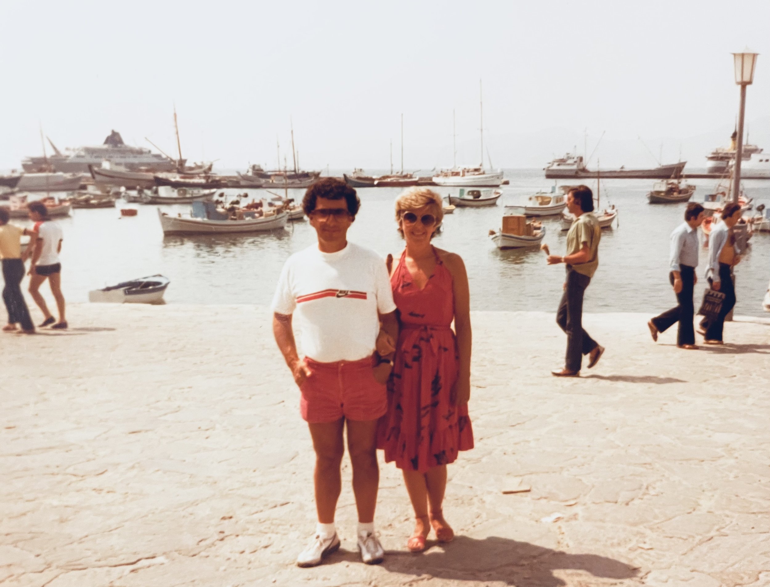 A vintage photo of two women standing on a stone pier by the water with boats and yachts docked in the background. They are smiling, one wearing a white t-shirt and red shorts, the other in a red dress and sunglasses. Several people are walking along