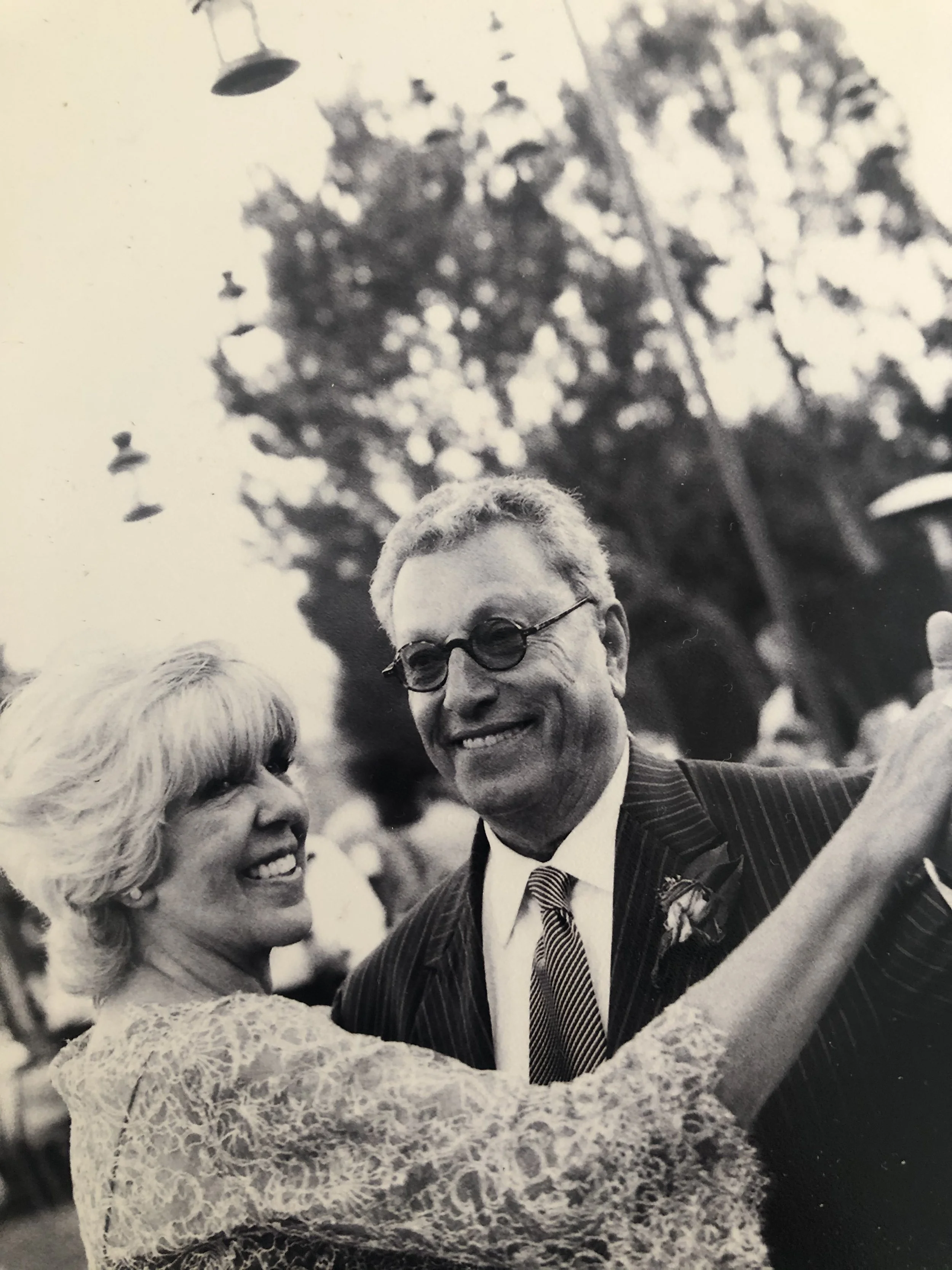 Black and white photo of an older couple dancing together outdoors during a celebration or event, with trees and hanging lights in the background.