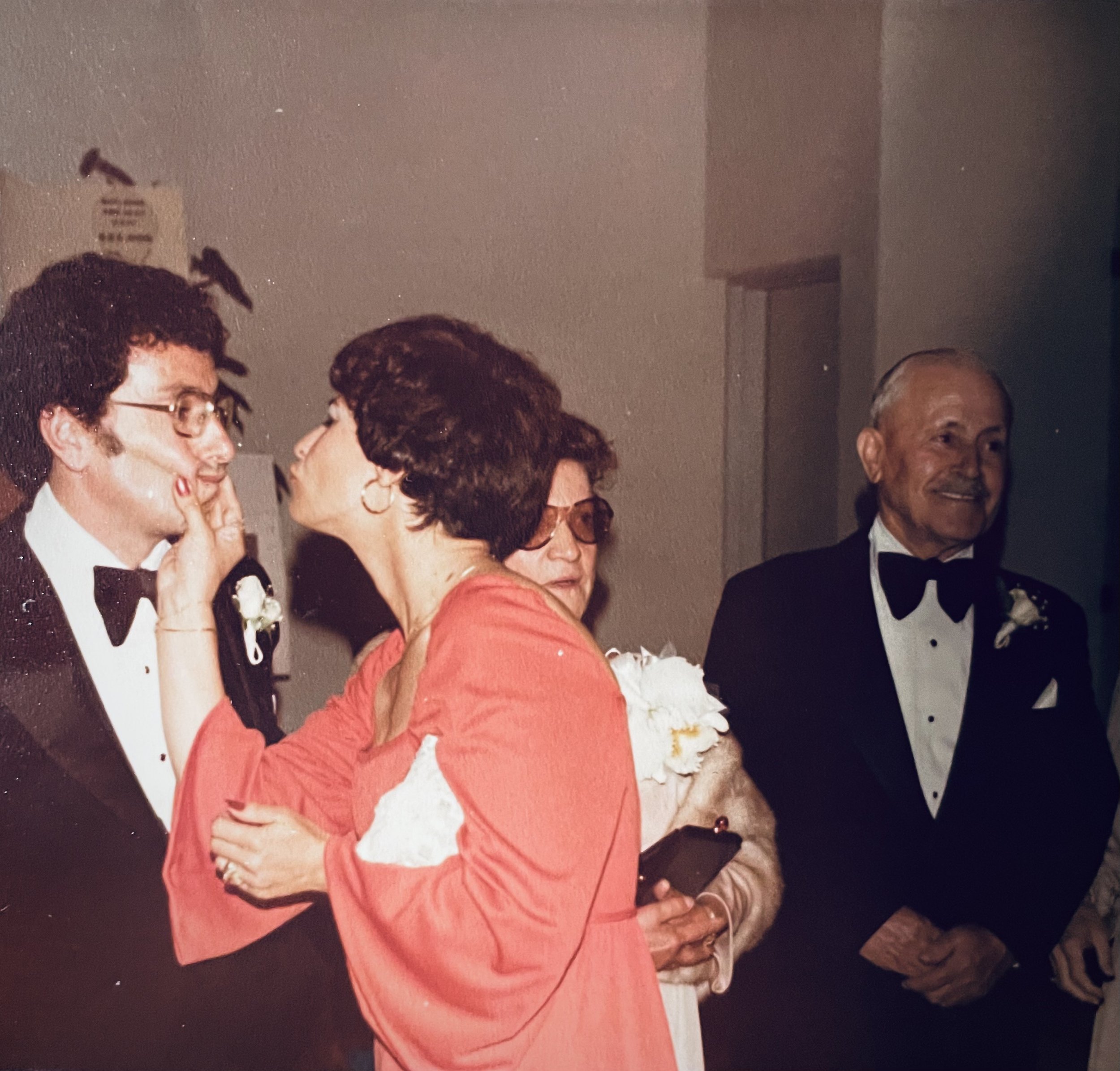 A vintage photograph of four people dressed in formal attire, possibly at a wedding or special event. The woman in the coral dress appears to be about to kiss the man in the tuxedo, who is wearing glasses and has dark hair. Two other women are observing, one with large sunglasses and light-colored clothing, and the man on the right is smiling, dressed in a tuxedo with a boutonniere.