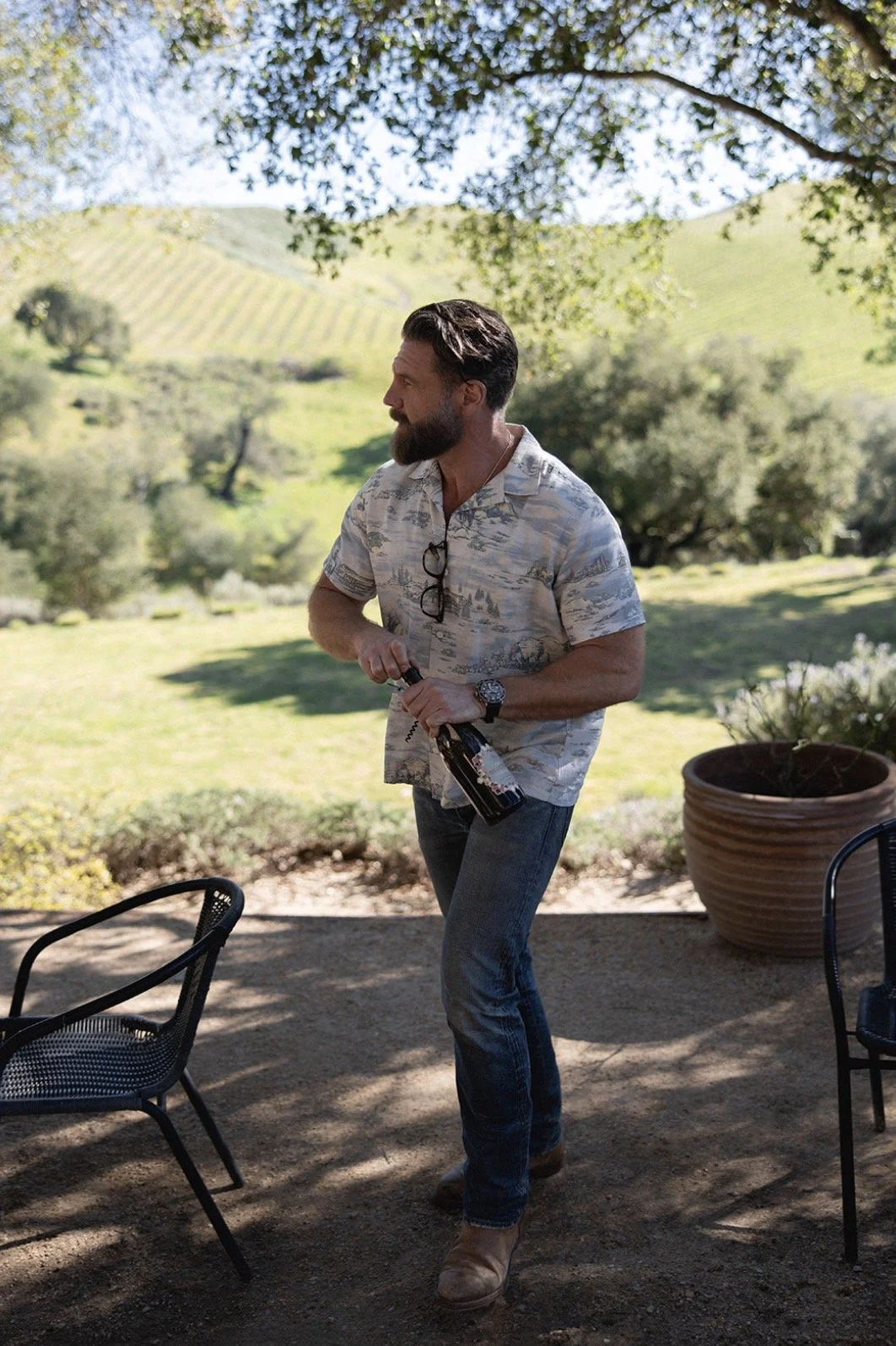 A man with a beard and glasses hanging on his shirt, holding a beer bottle, standing outdoors under a shade tree with a scenic hillside background, during daytime.
