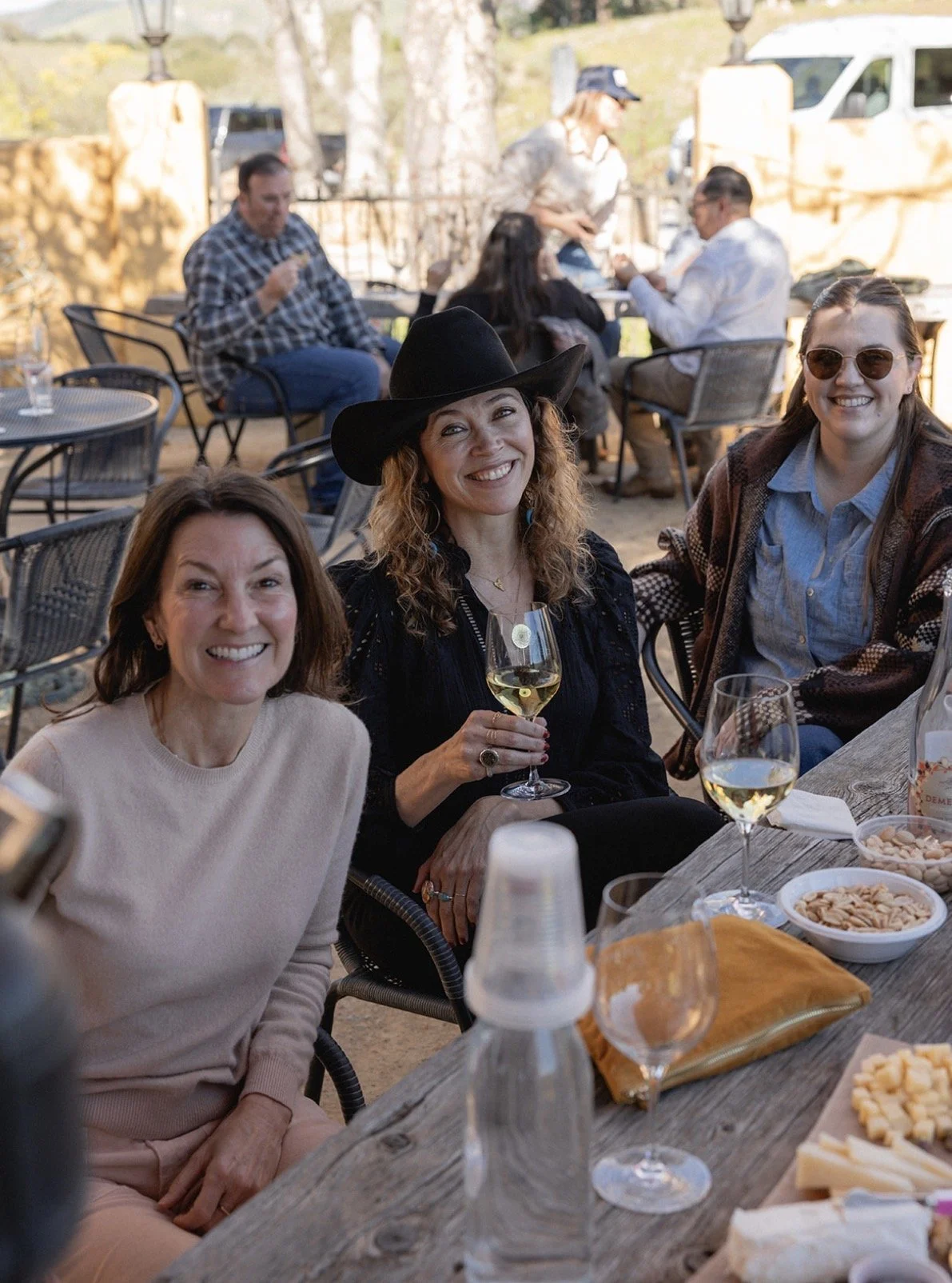 A group of women enjoying a wine at an outdoor gathering, with others in the background engaged in conversation, on a rustic patio with trees and outdoor furniture.