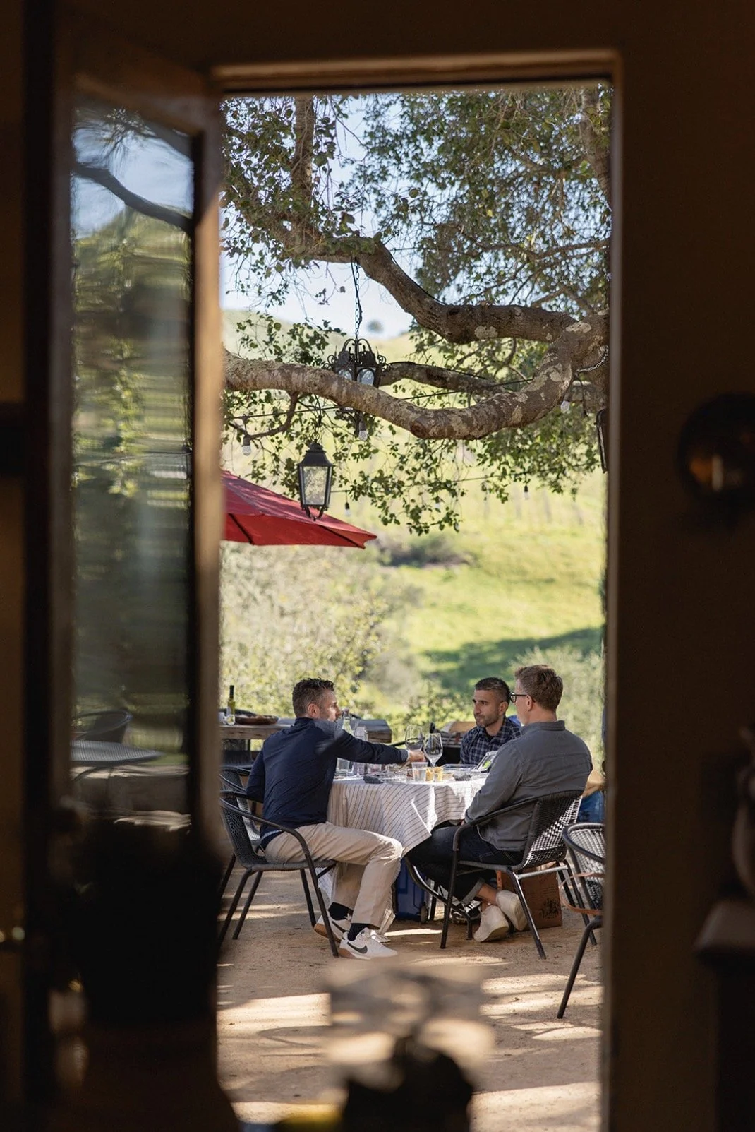 Three men dining outdoors at a round table with a white tablecloth, set with glasses and dishes, under a large tree with hanging lanterns and a red umbrella in the background.