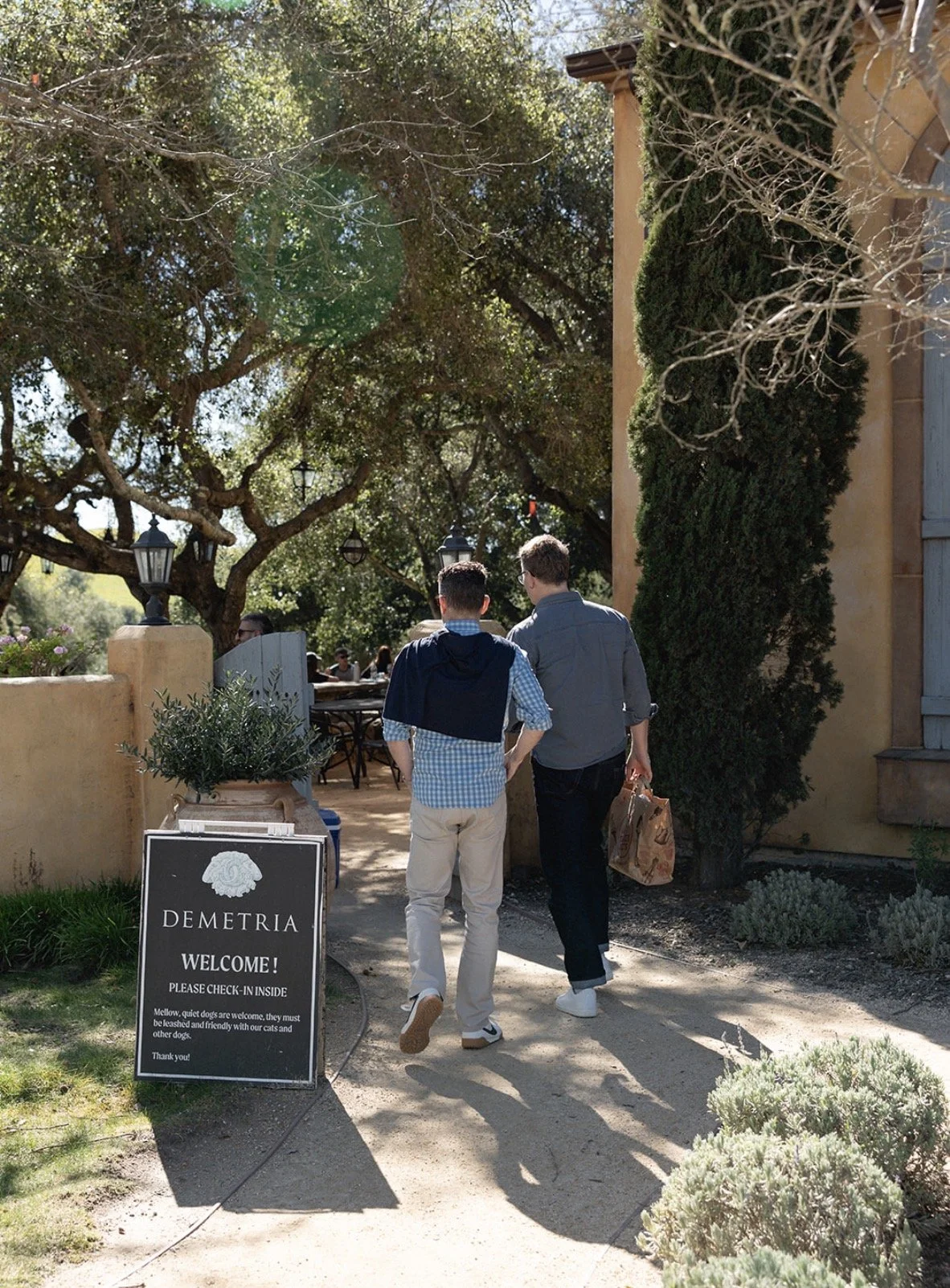 Two men walking on a garden pathway toward a restaurant entrance under trees, with a signboard reading 'Demetria. Welcome! Please check in inside.'