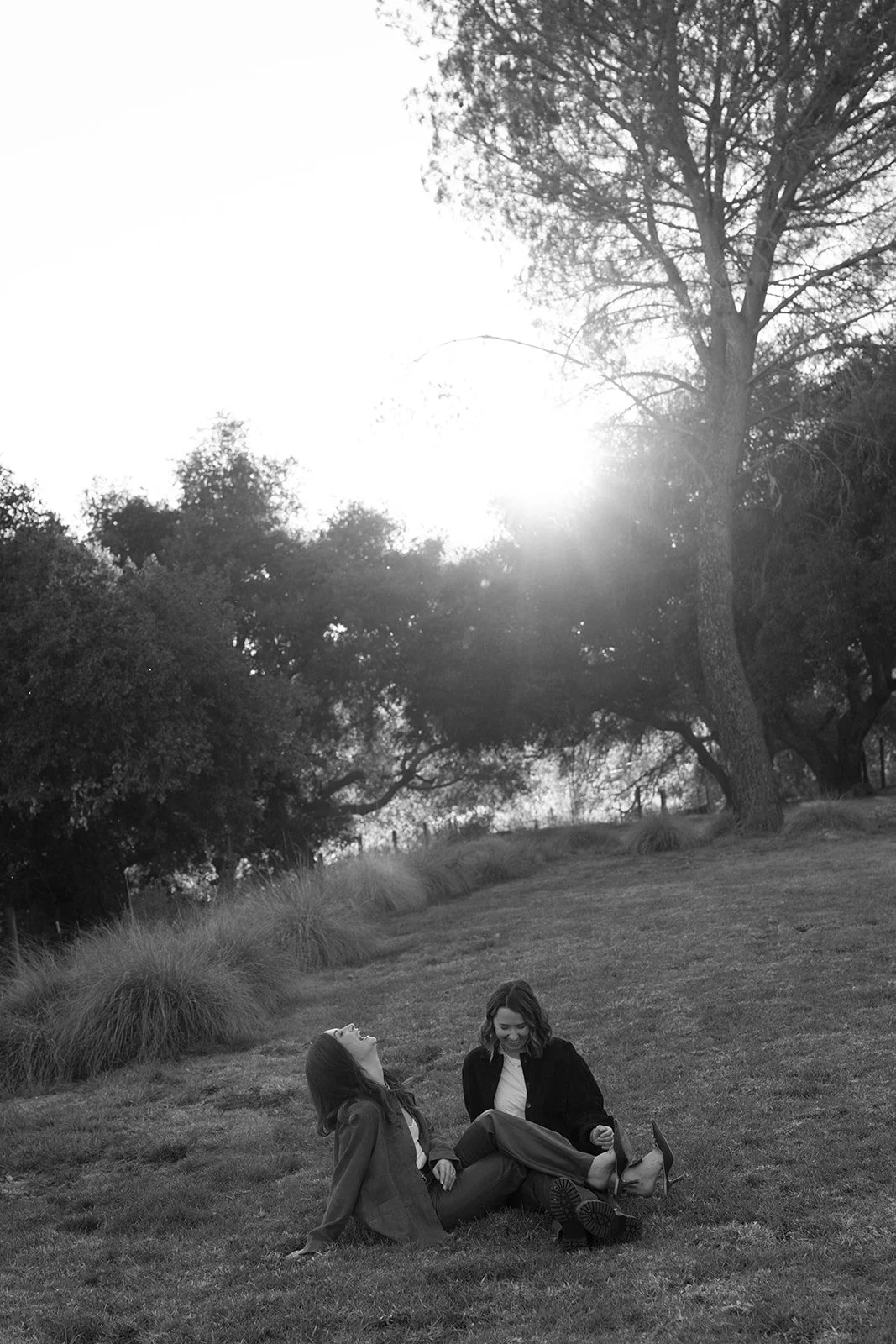 Two women sitting on grass in a park, laughing and smiling, with trees and sunlight in the background.