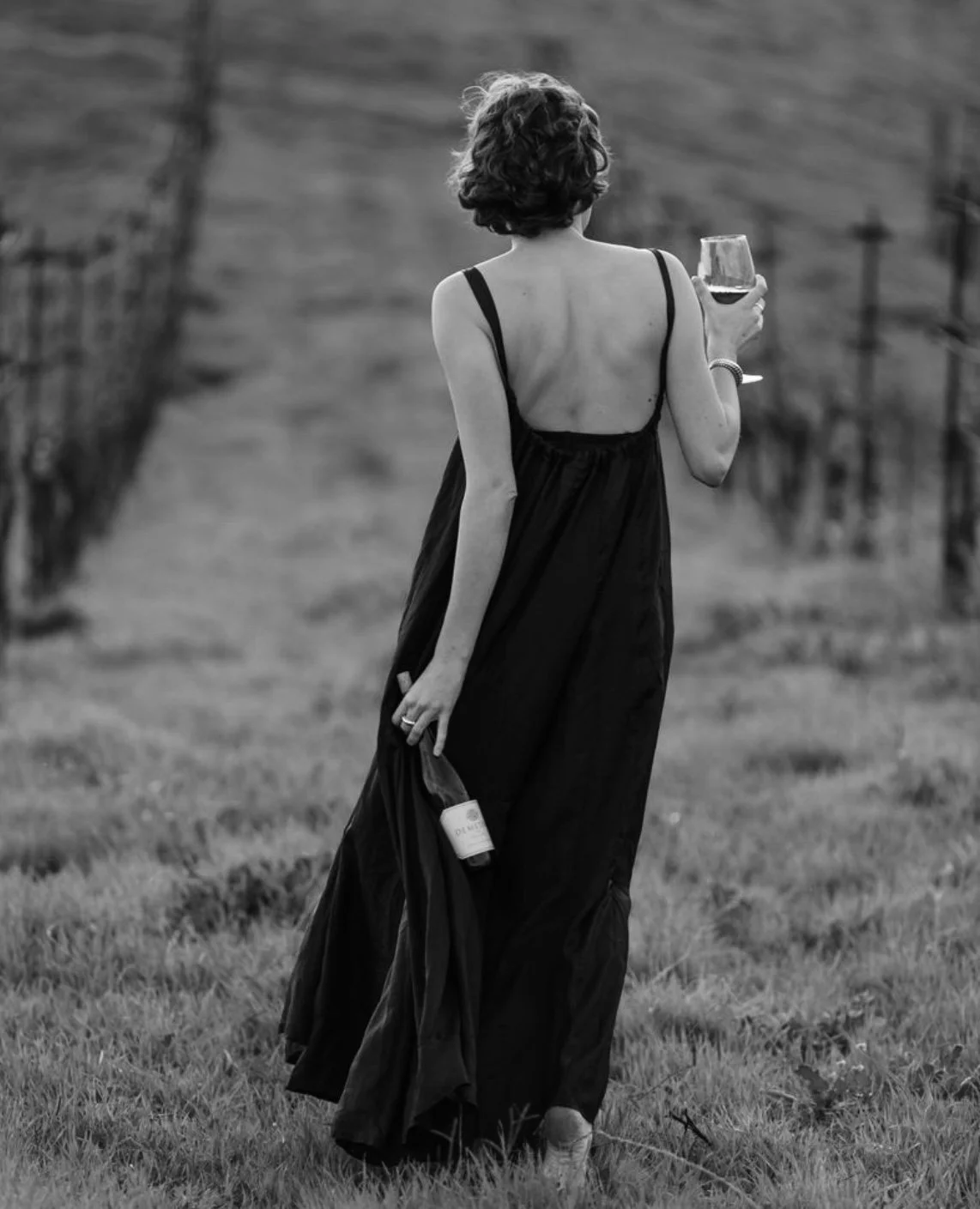 A woman in a long, backless dress holding a wine glass and a bottle of wine while walking through a vineyard.