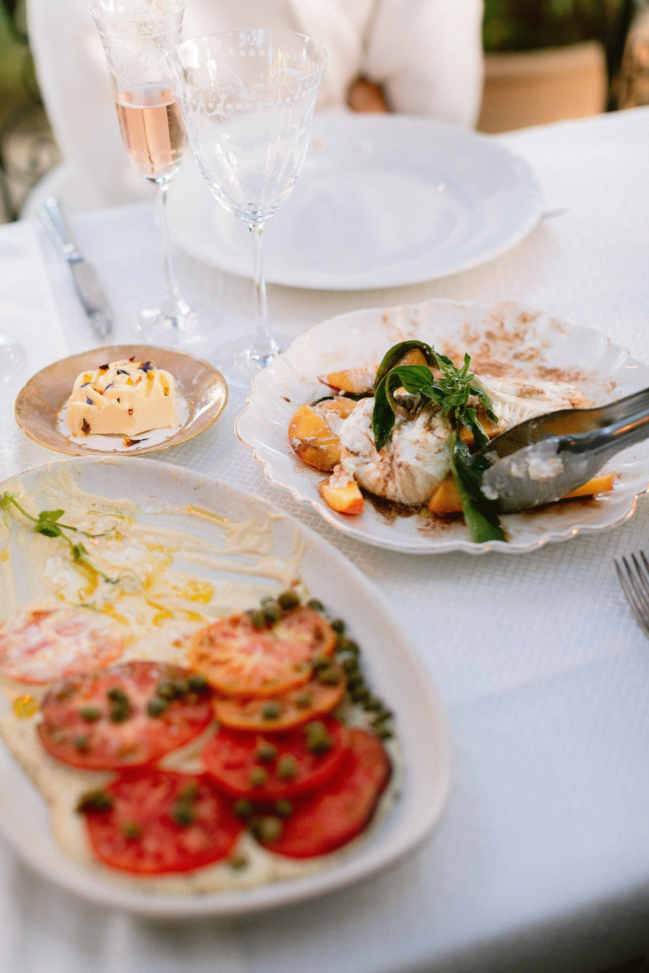 Elegant dining table set with plates of food, including a fish dish with greens and vegetables, a sliced tomato and potato dish, and butter with herbs, along with glasses of rosé wine and champagne.