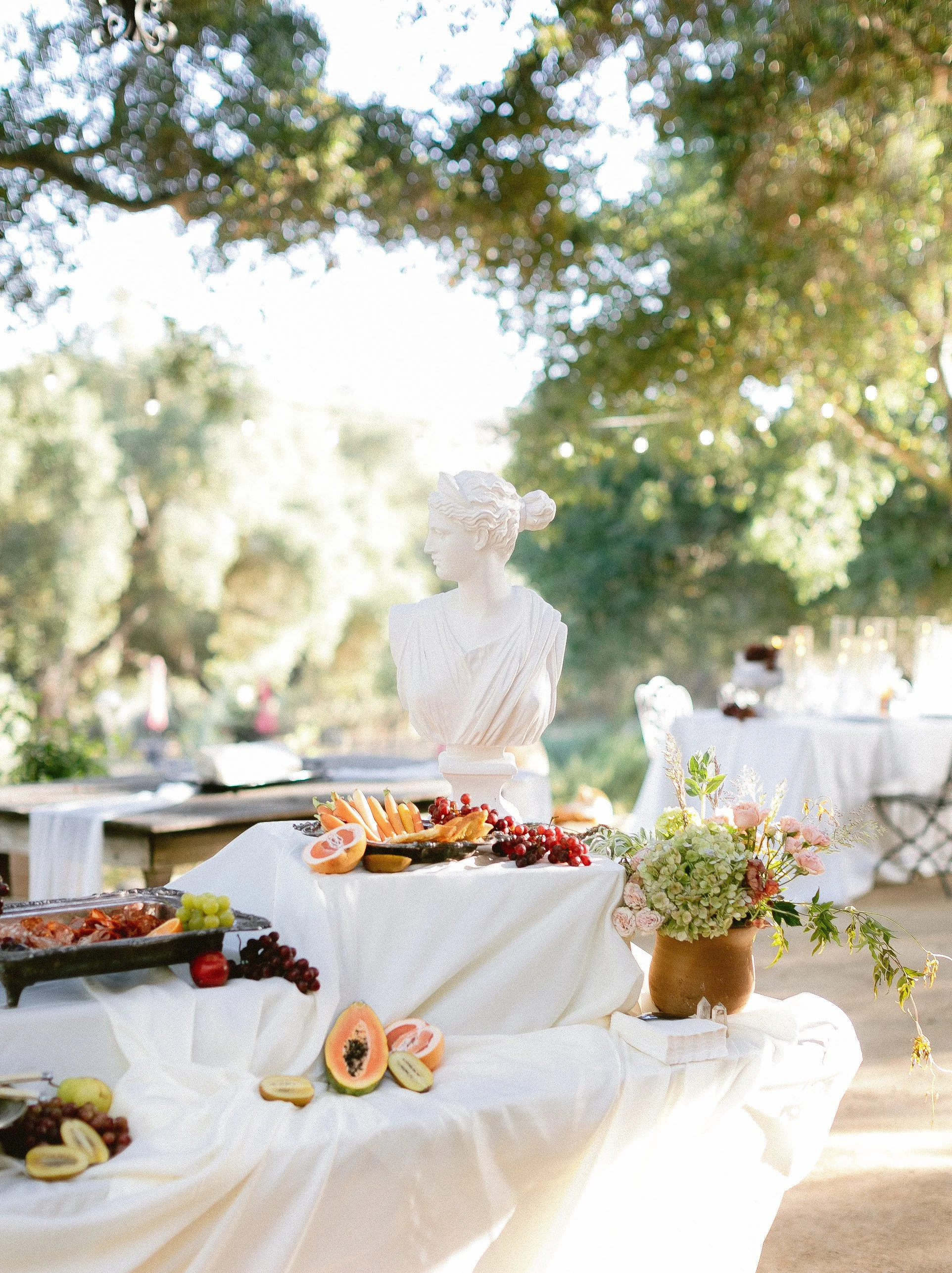 Outdoor table decorated with a white cloth, flowers in a brown vase, and a bust statue surrounded by an assortment of fresh fruit including grapes, papaya, and oranges, in a garden setting with trees and string lights.