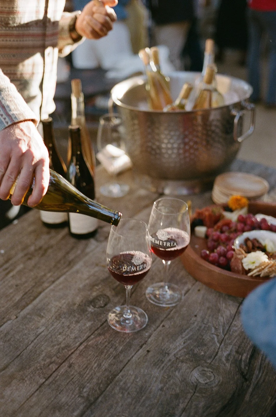 Person pouring red wine into a glass at a rustic outdoor gathering, with a bowl of grapes and cheese on the table.