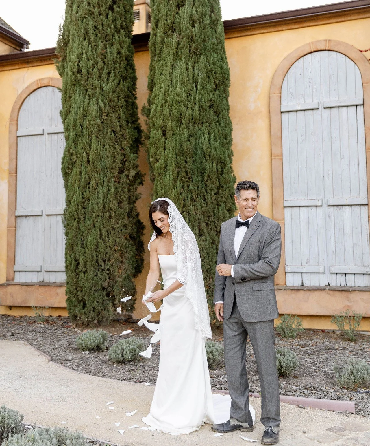 Bride in white wedding dress and veil smiling and throwing flower petals, standing next to an older man in gray suit and bowtie outside near tall cypress trees and a yellow building with blue wooden shutters.