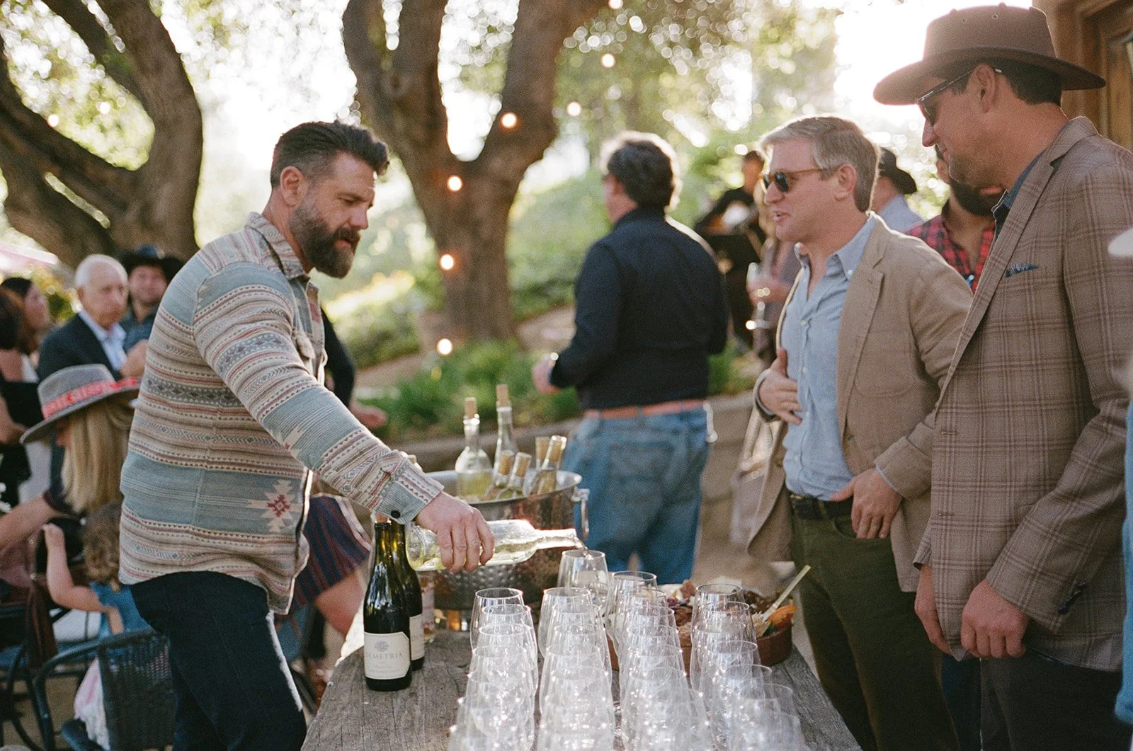 Man pouring wine at outdoor gathering with people socializing and a large tree in the background.