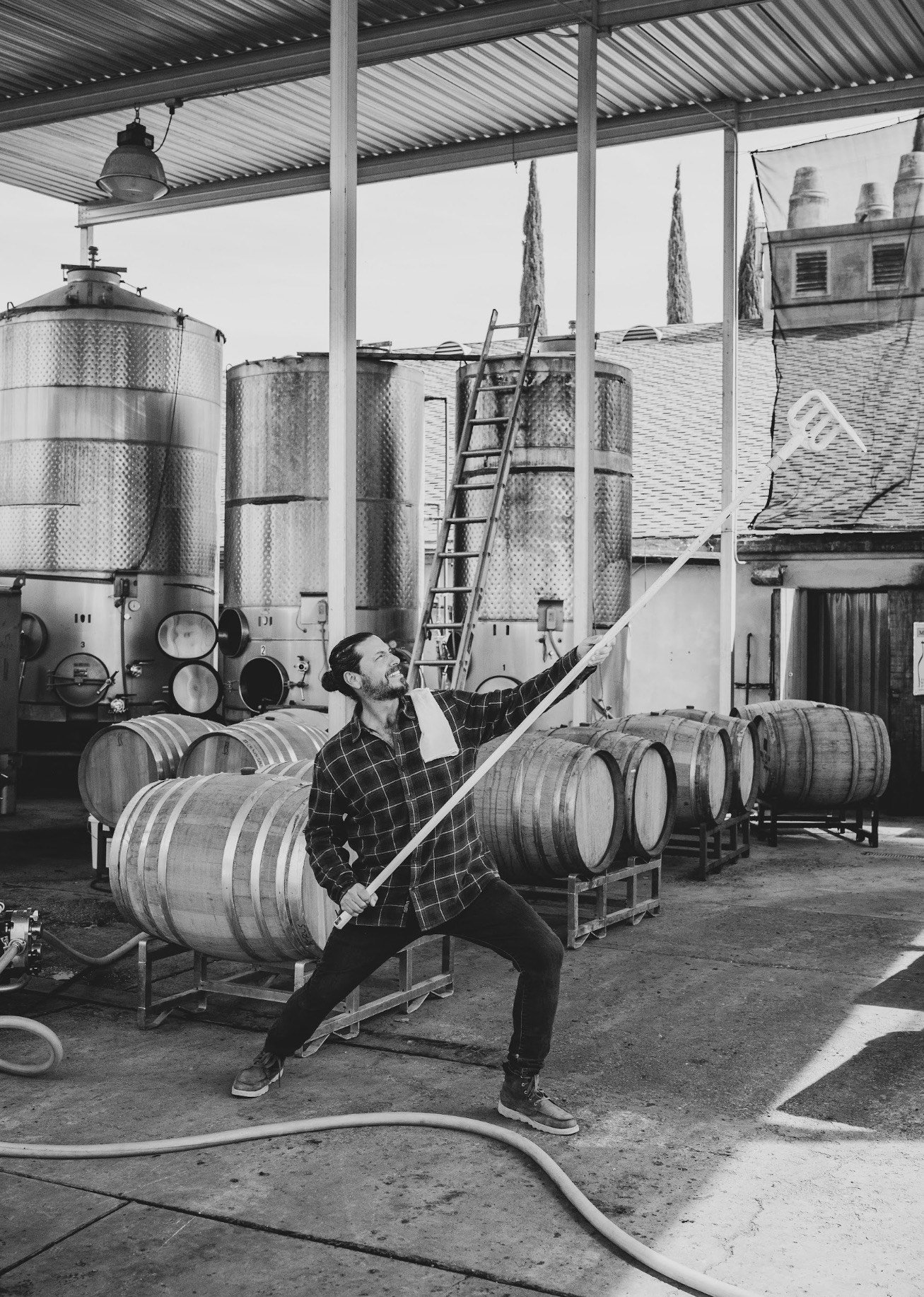 A man is standing in a winery or brewery, holding a long pole and smiling, surrounded by large metal tanks and wooden barrels.
