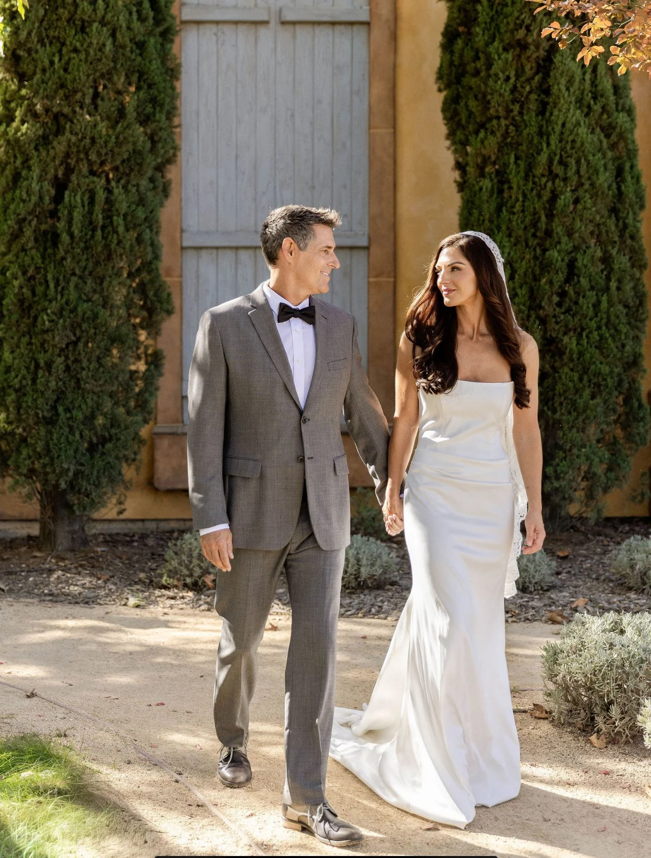 A couple, dressed in wedding attire, walking hand in hand outdoors on a sunny day.