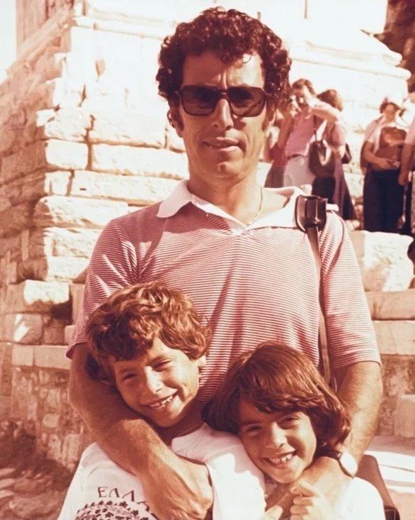 A man with curly dark hair, wearing sunglasses and a striped polo shirt, stands on stone stairs with two young children, a boy and a girl, both smiling and leaning into him. The background shows other people on the stairs.