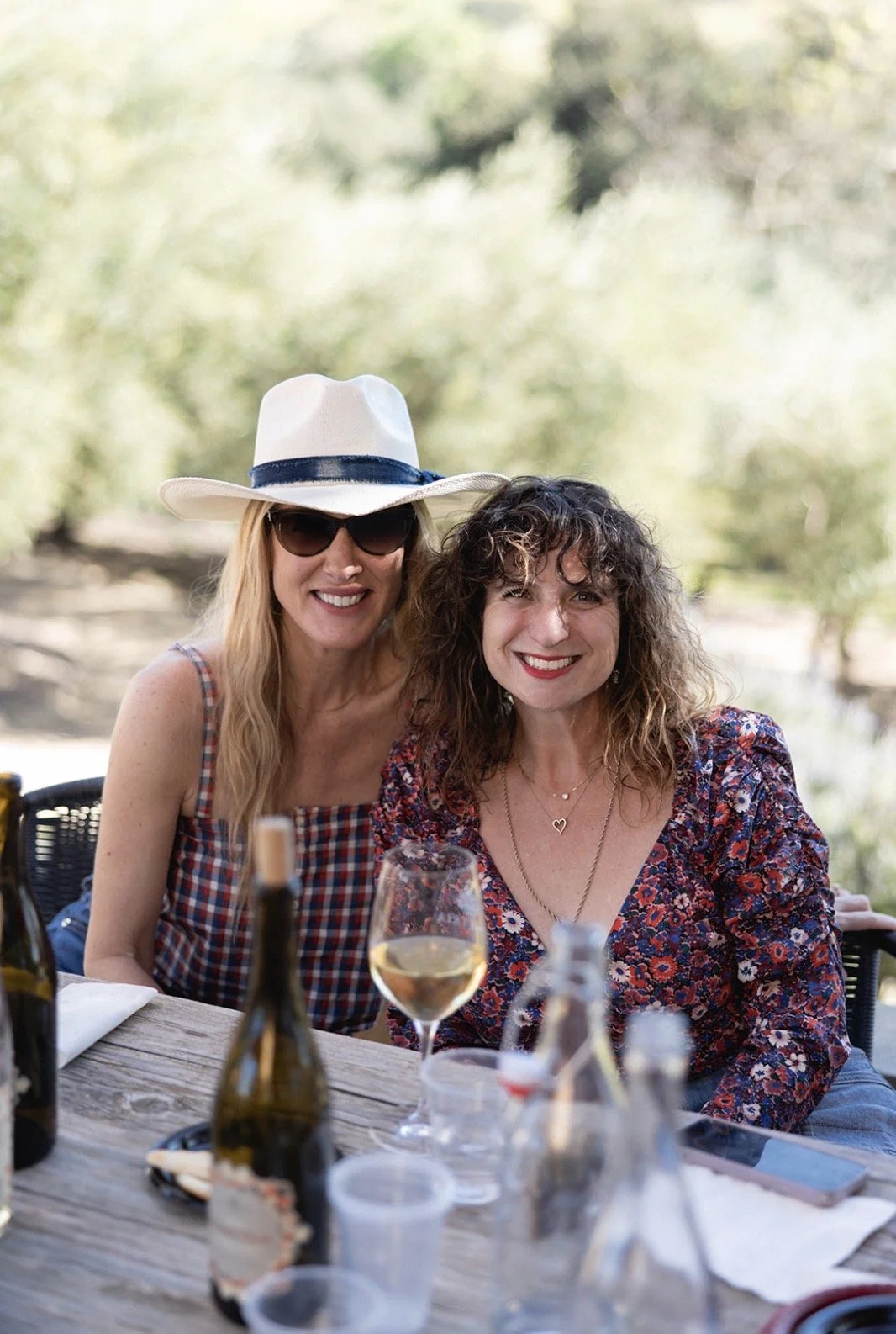Two women smiling at an outdoor dining table with wine bottles and glasses, surrounded by blurred greenery.