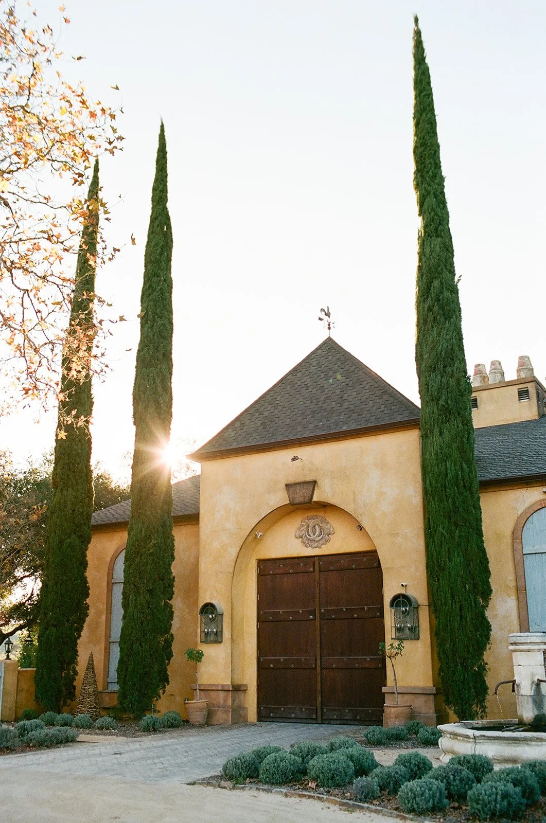 A large house with a yellow stucco facade, dark wooden double doors, three small trees on each side, tall cypress trees, and a stone fountain in the front yard, with the sun setting behind the house.