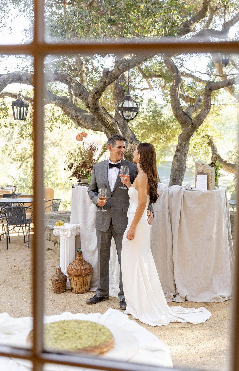 A bride and groom holding wine glasses sharing a moment at their wedding reception outdoors with trees and lanterns in the background.
