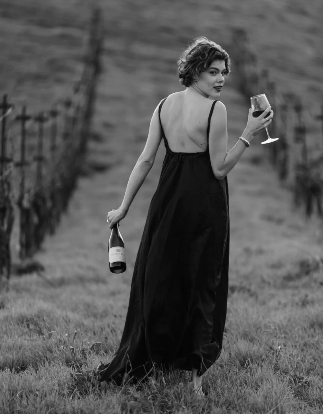 A woman in a black dress holding a glass of wine and a bottle of wine walking through a vineyard.