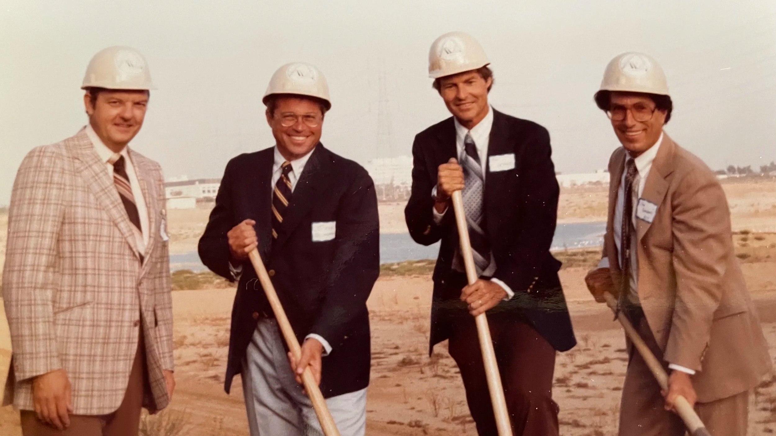 Four men wearing suits and construction helmets on a dirt site with a river and buildings in the background, holding shovels.