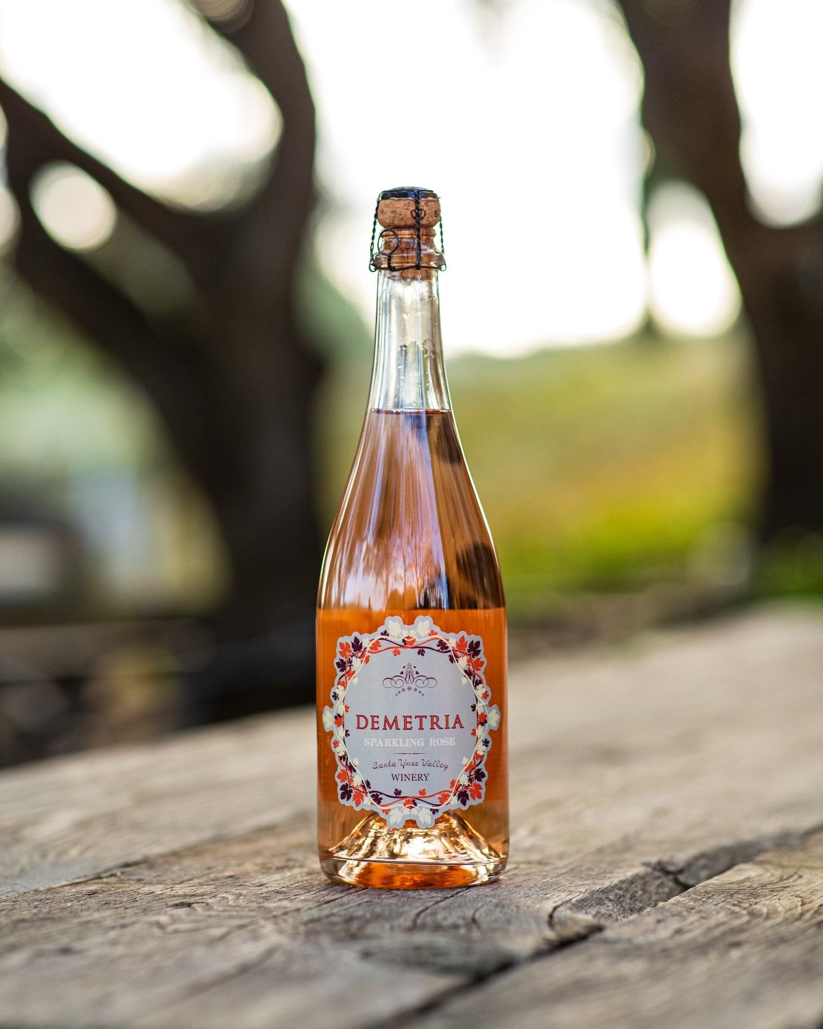 A bottle of sparkling rosé wine with a cork and wire cage, placed on a wooden surface outdoors with trees in the background.