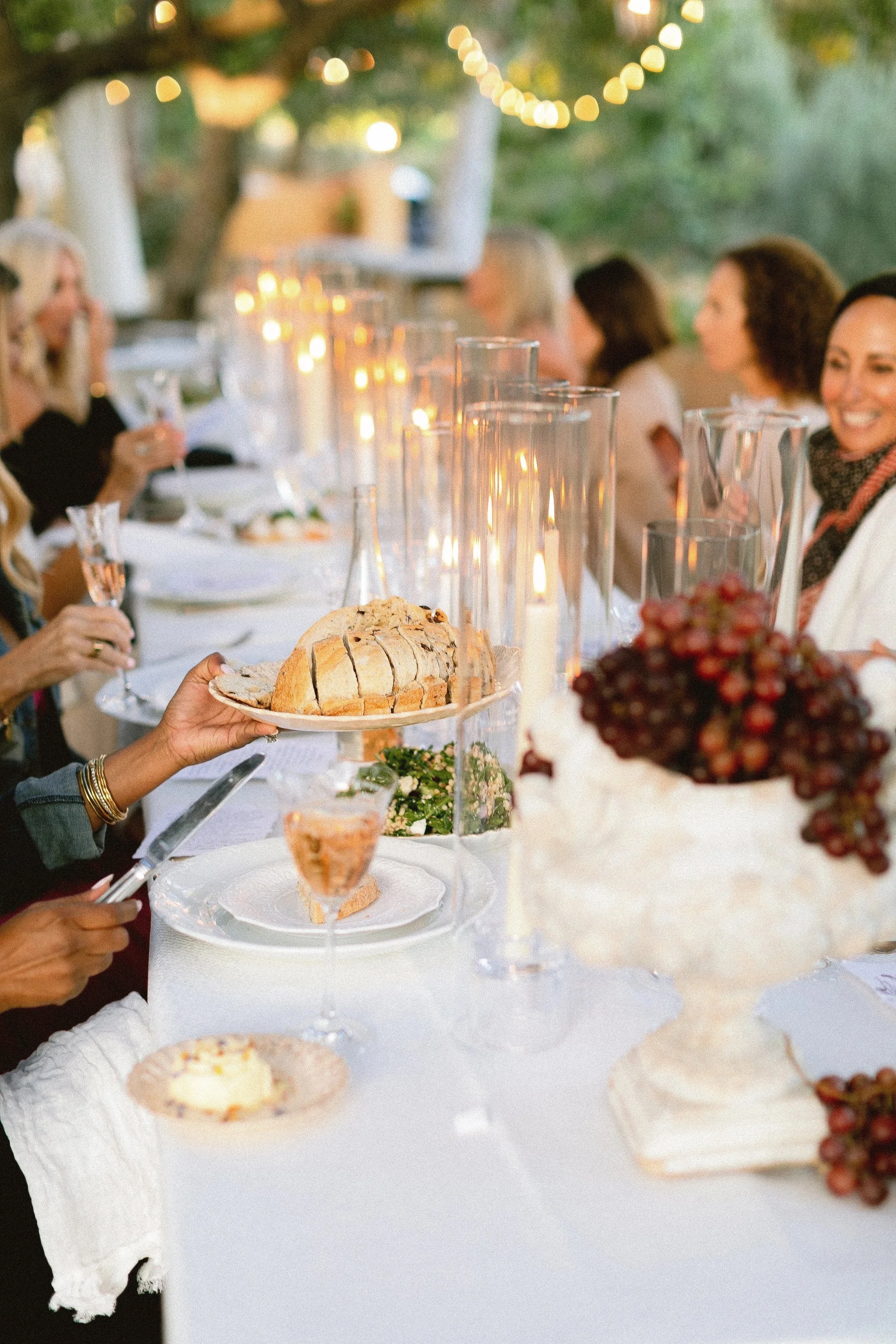 People gathered at an outdoor dinner party with string lights, a long table with white tablecloth, glasses of wine, a loaf of sliced bread, a bowl of grapes, and candles.