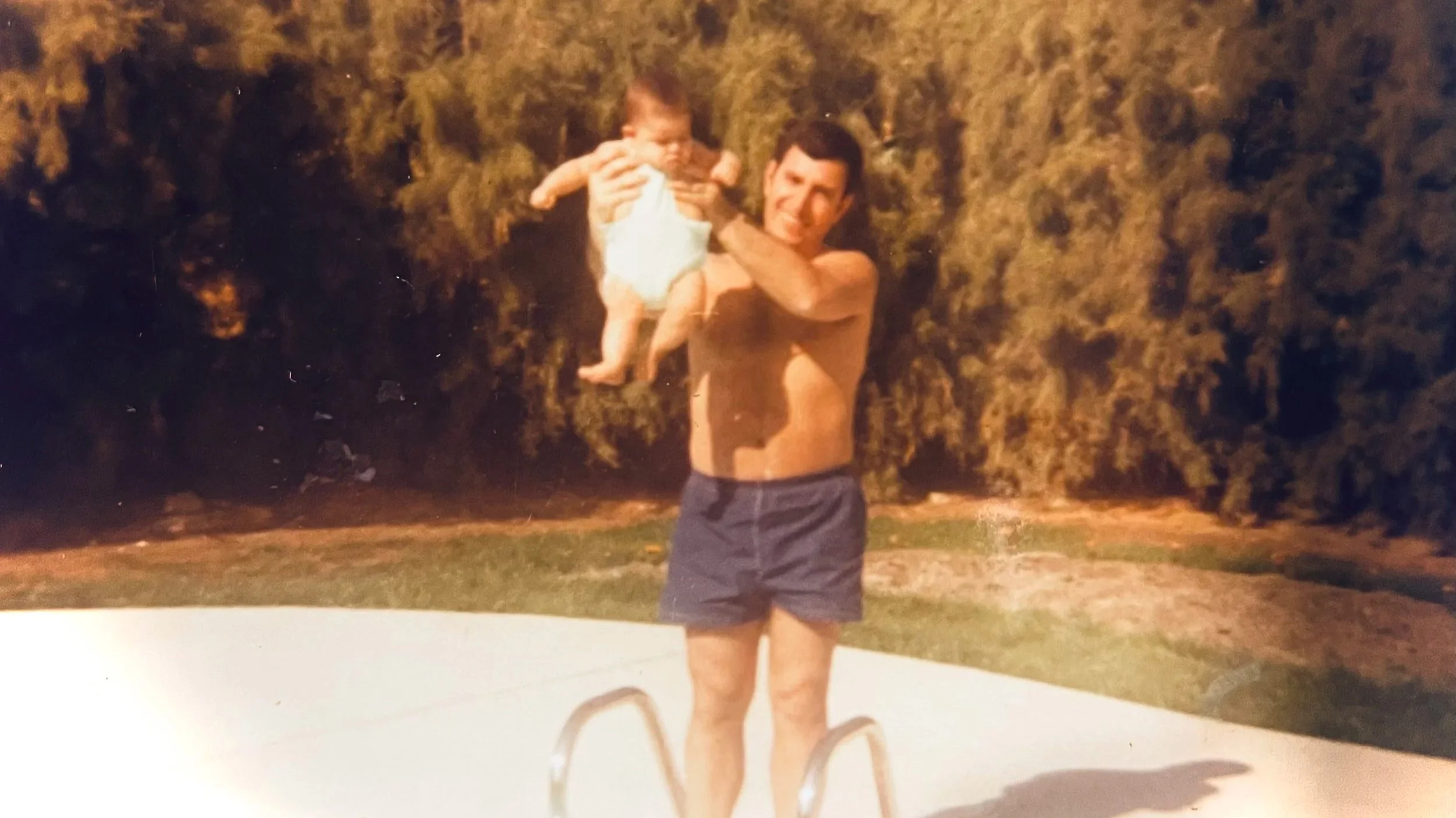 A man holding a baby near a swimming pool in an outdoor setting with trees in the background.