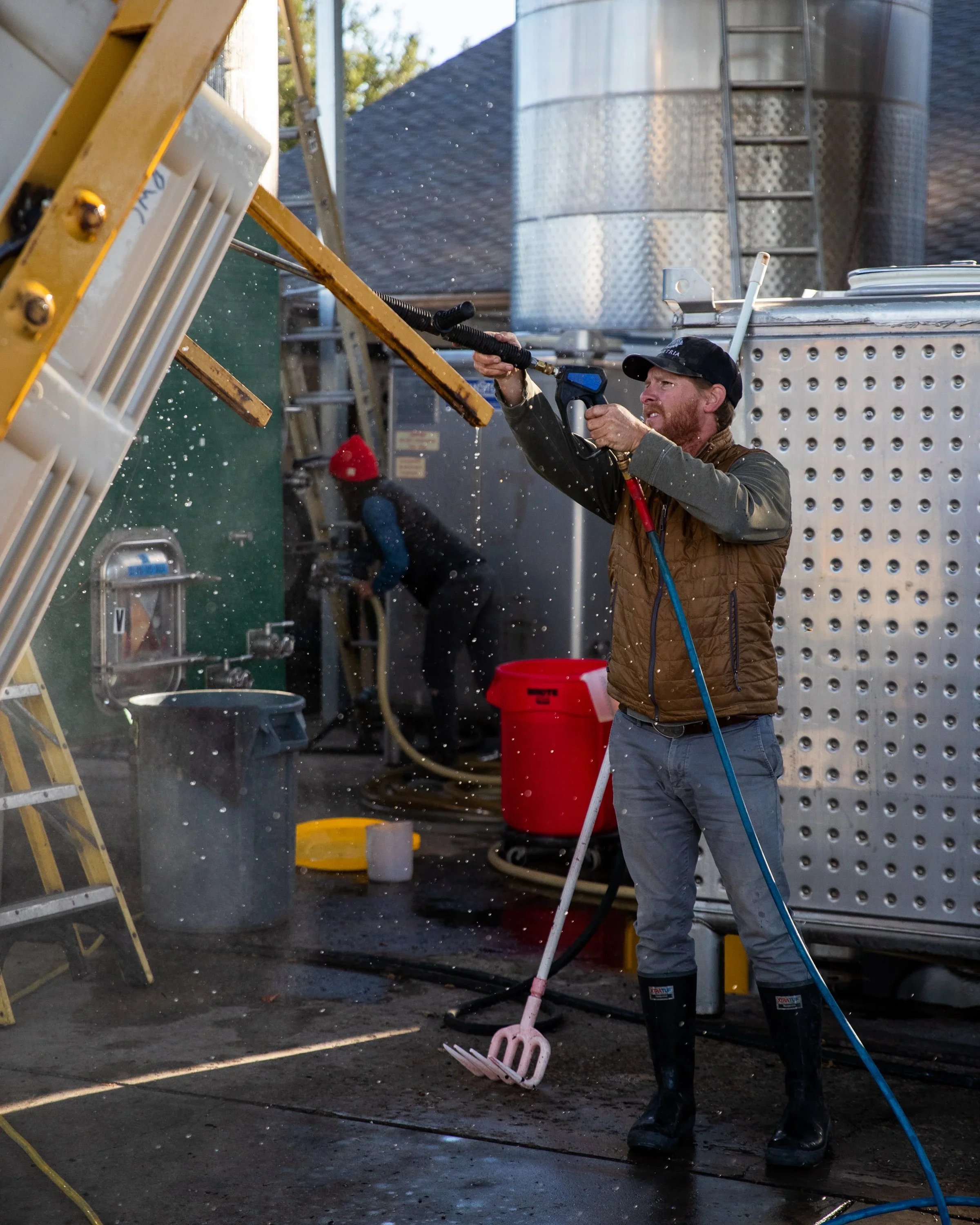 A man wearing a jacket and boots is power washing a metal surface outdoors. Another person in the background is working near large tanks, and there is cleaning equipment and hoses around.