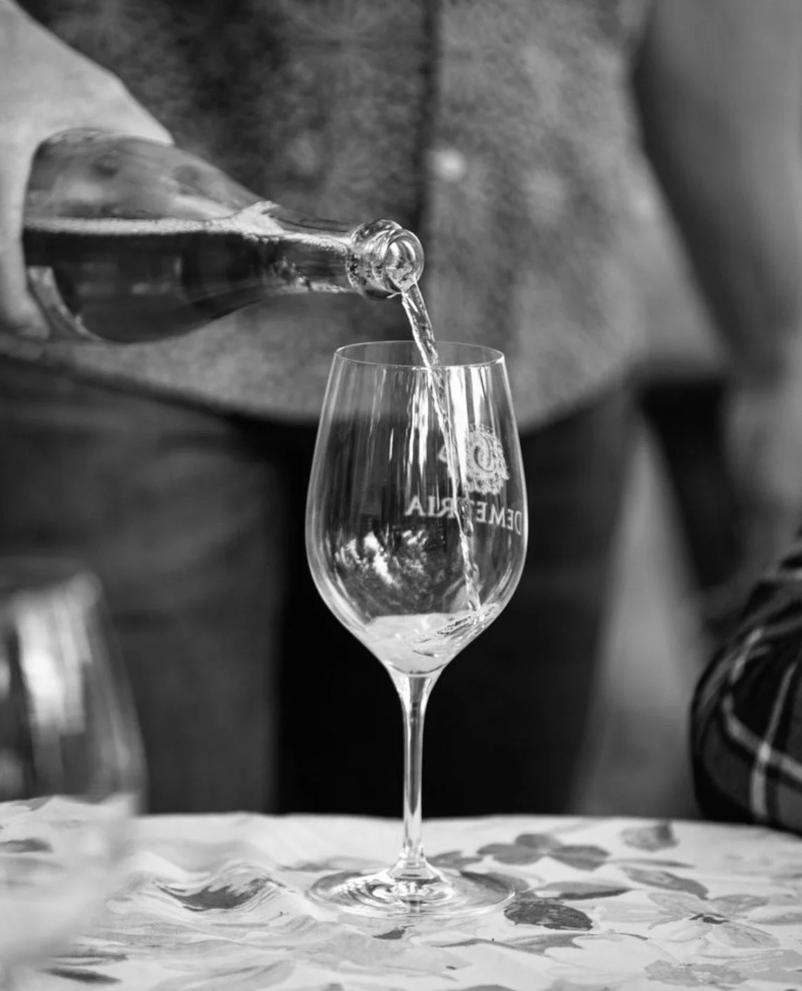 A person is pouring a drink into a Stemmed glass with an engraved logo on it, on a tablecloth with a floral pattern.