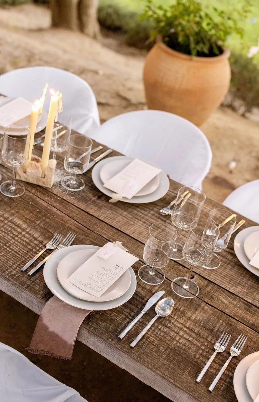 A rustic wooden dining table set for a meal with white plates, silverware, wine glasses, and a menu. The table has a beige table runner and white chairs with cloth covers. In the center, there's a candles holder with two lit candles. In the backgroun