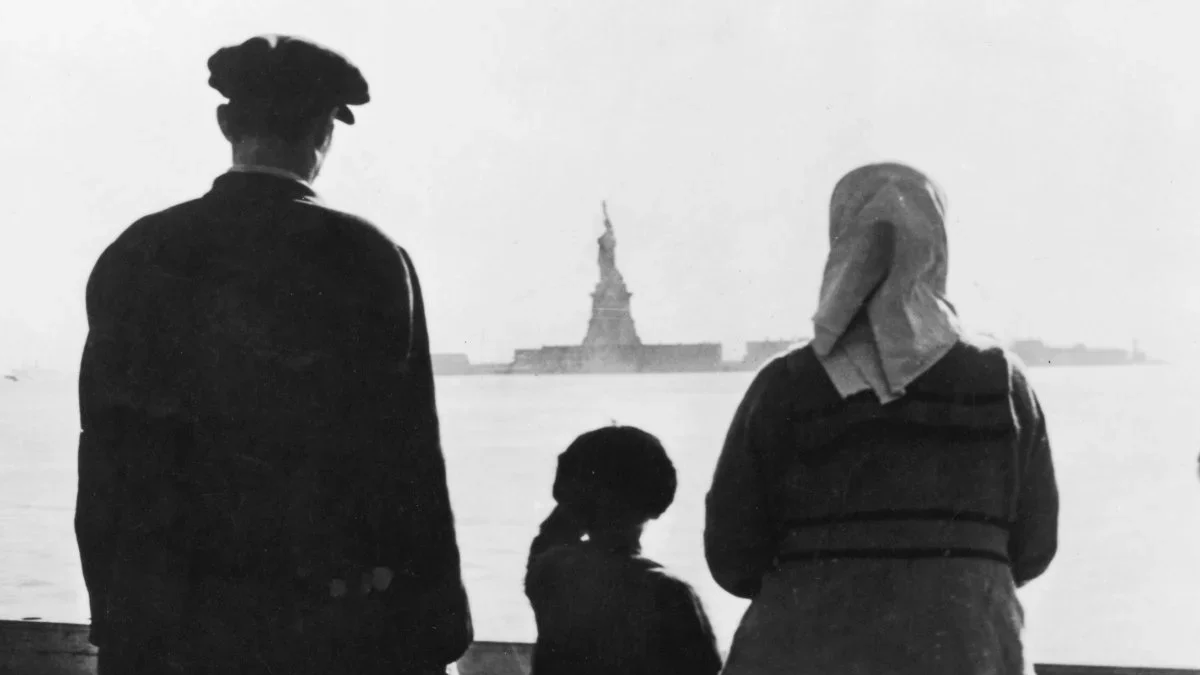 Three people standing on a shoreline, observing the Statue of Liberty across the water.