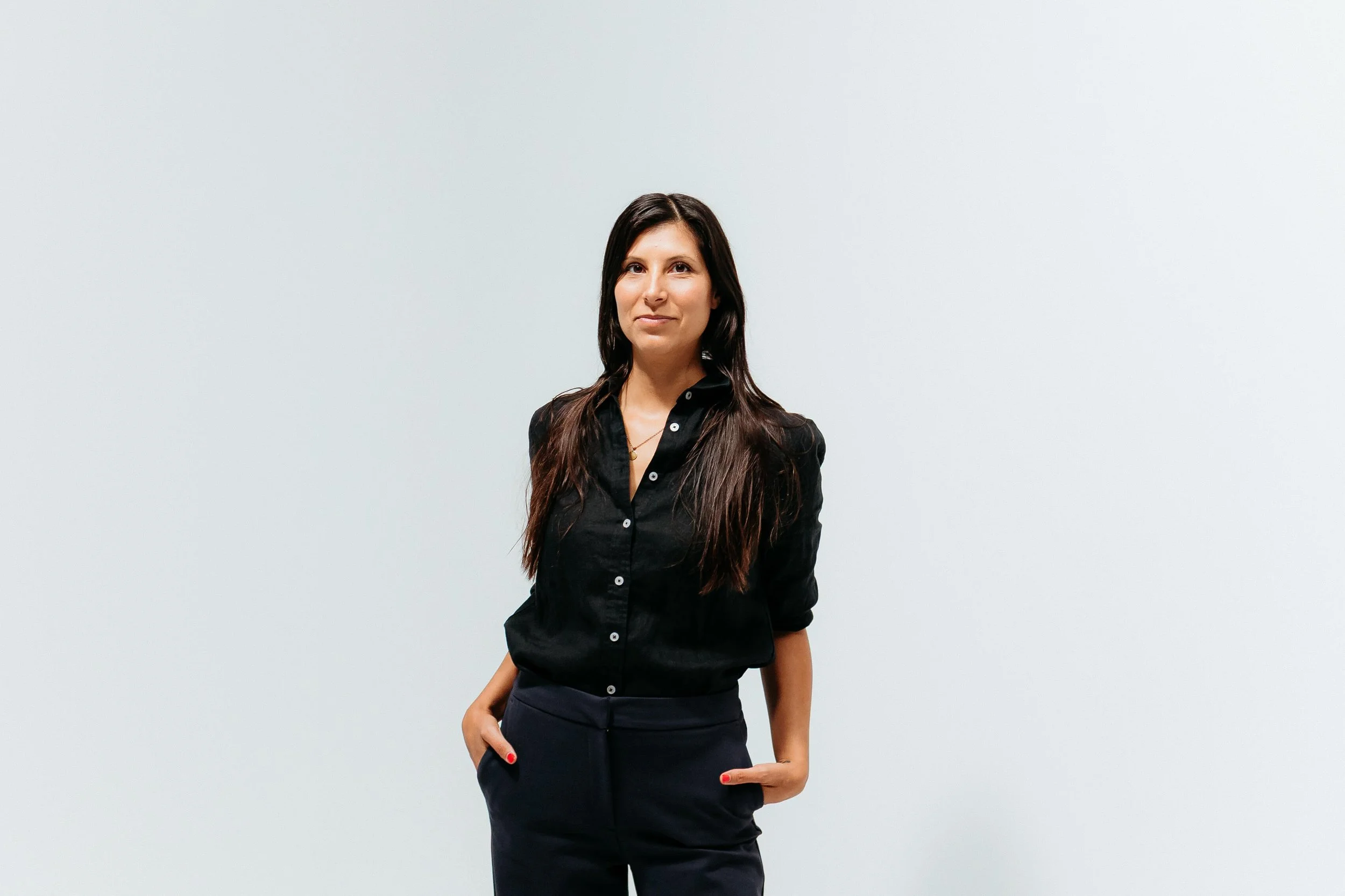 A woman with long dark hair wearing a black button-up shirt and navy pants standing against a plain light grey wall.