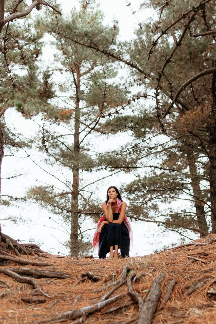 A woman sitting on the ground in a forest, surrounded by pine trees, with her chin resting on her hand, looking thoughtful.