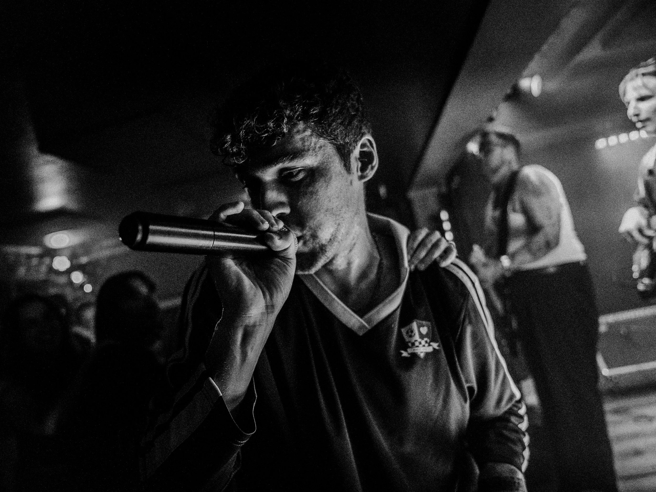 A young man holds a microphone close to his mouth in a black and white photo, with a blurred crowd and band members in the background.