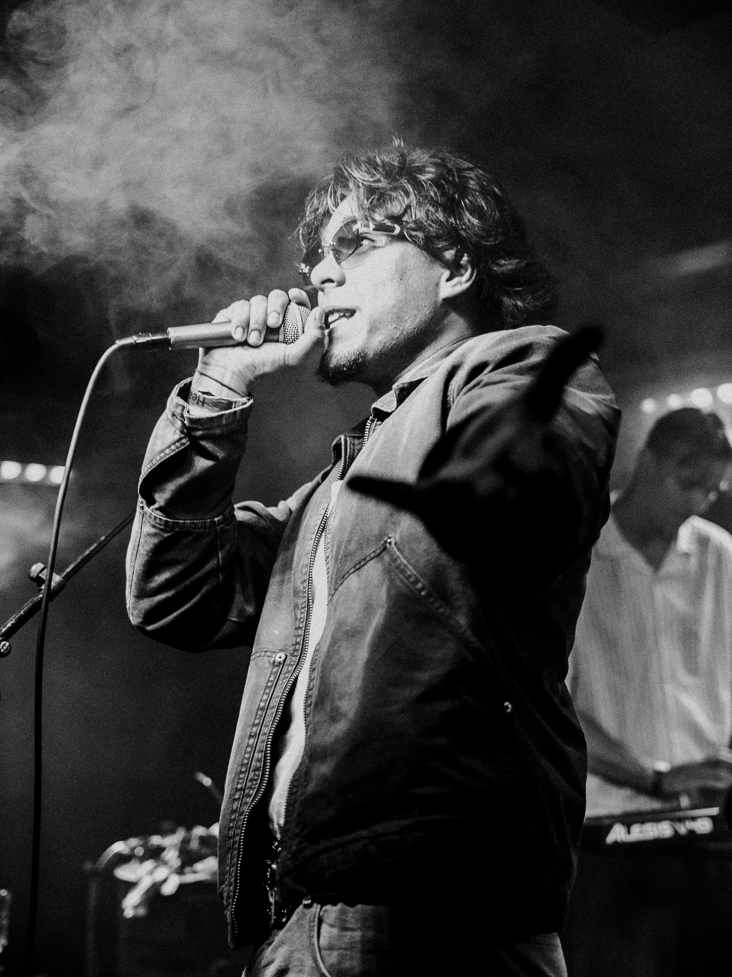 A man with curly hair and sunglasses singing into a microphone on stage, wearing a jacket, with a keyboard player in the background.