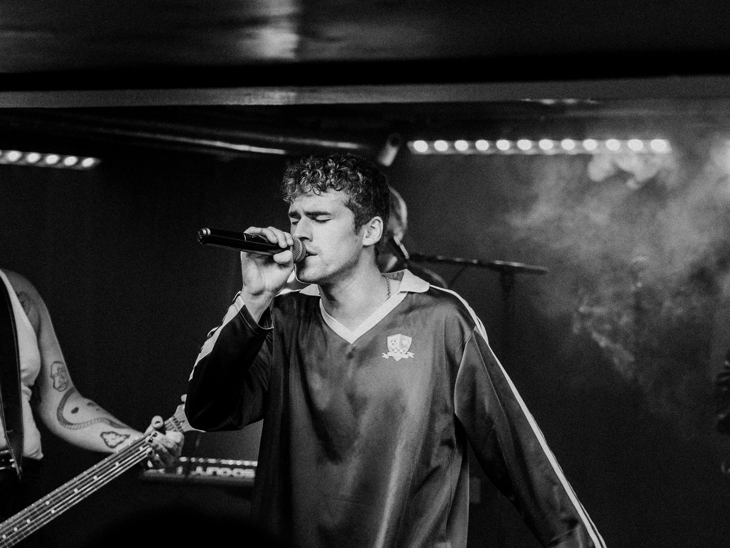 A male singer with curly hair passionately singing into a microphone on stage, with a guitarist in the background.