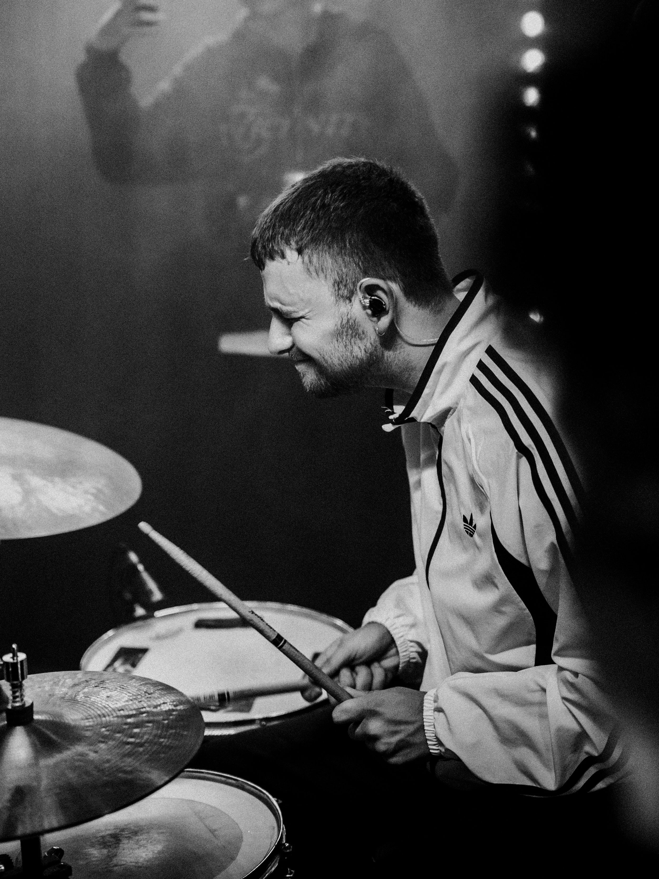 A man playing drums, smiling and focused, wearing a white Adidas jacket with black stripes, in a dark setting.