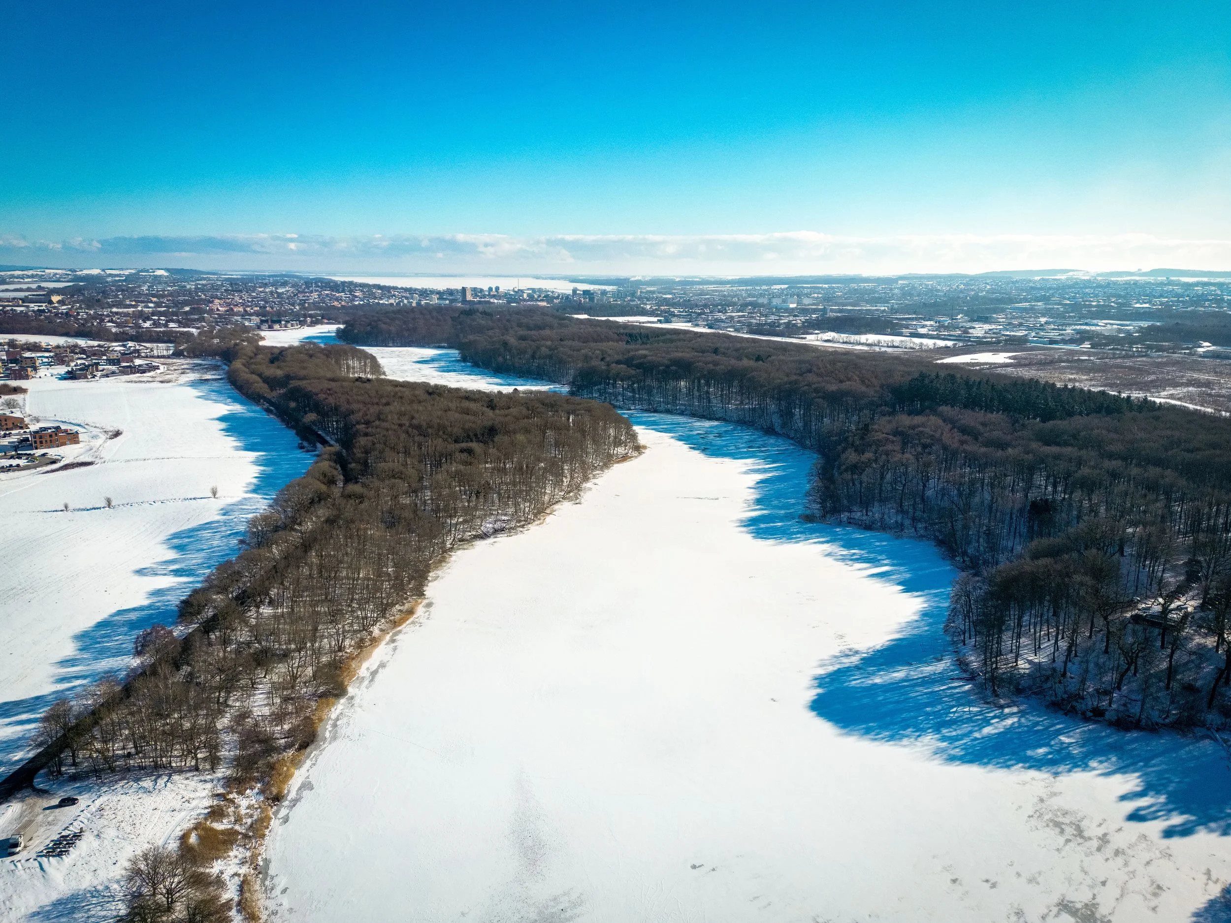 Aerial view of a snowy river surrounded by leafless trees and open fields on a sunny winter day.