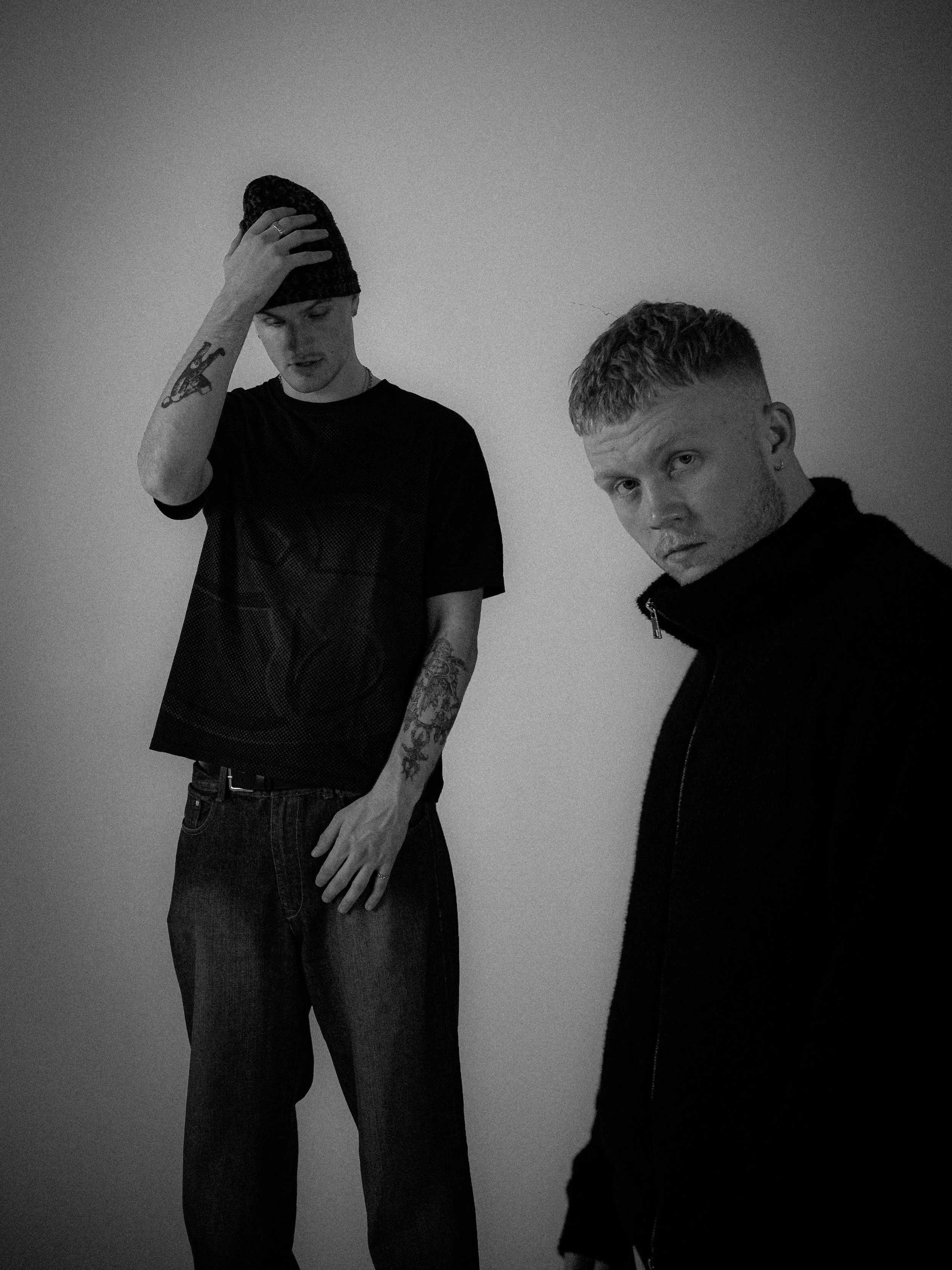 Two young men are standing against a plain wall, one touching his forehead and the other looking intently at the camera, all in black and white.