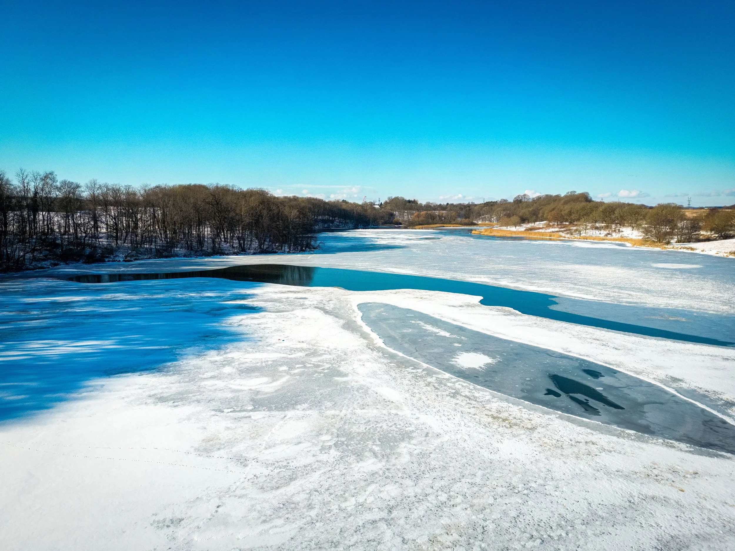 Frozen river with patches of ice and snow, surrounded by trees and a clear blue sky.