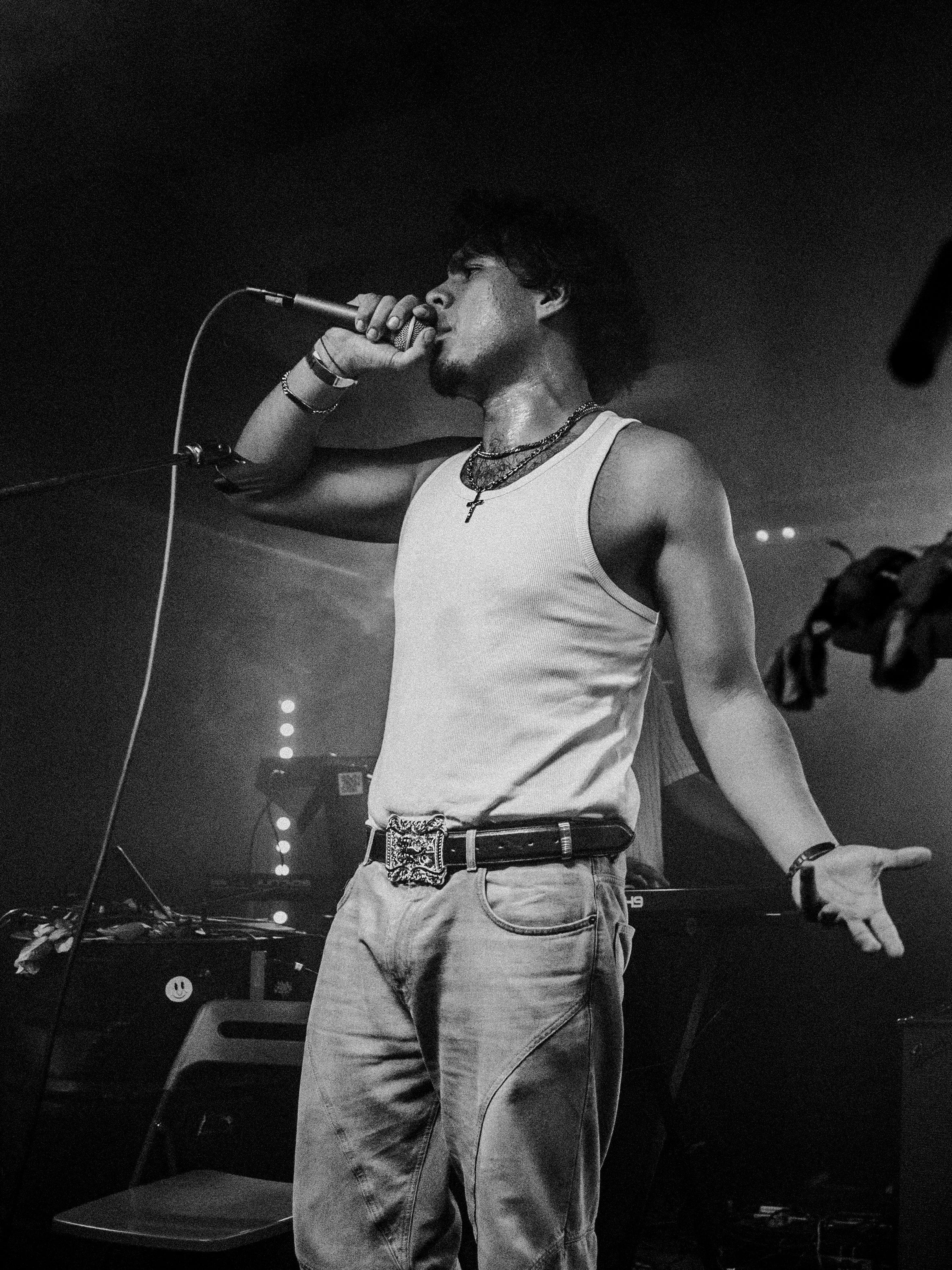 A male singer during a performance, holding a microphone and wearing a tank top, necklace, and belt, on a dimly lit stage.